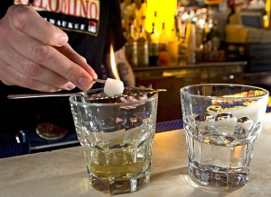 The preparation of absinthe requires running ice water over a sugar cube in a slotted spoon and into the yellow liquid itself.