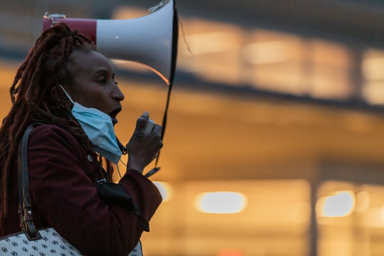 A woman with her mask hanging on one ear speaks loudly into a megaphone, which she holds next to her head