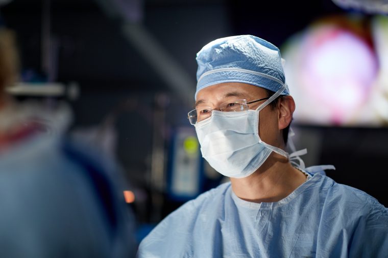 A man in a light blue medical mask and blue medical gear is shown in a medical lab with a blurry background