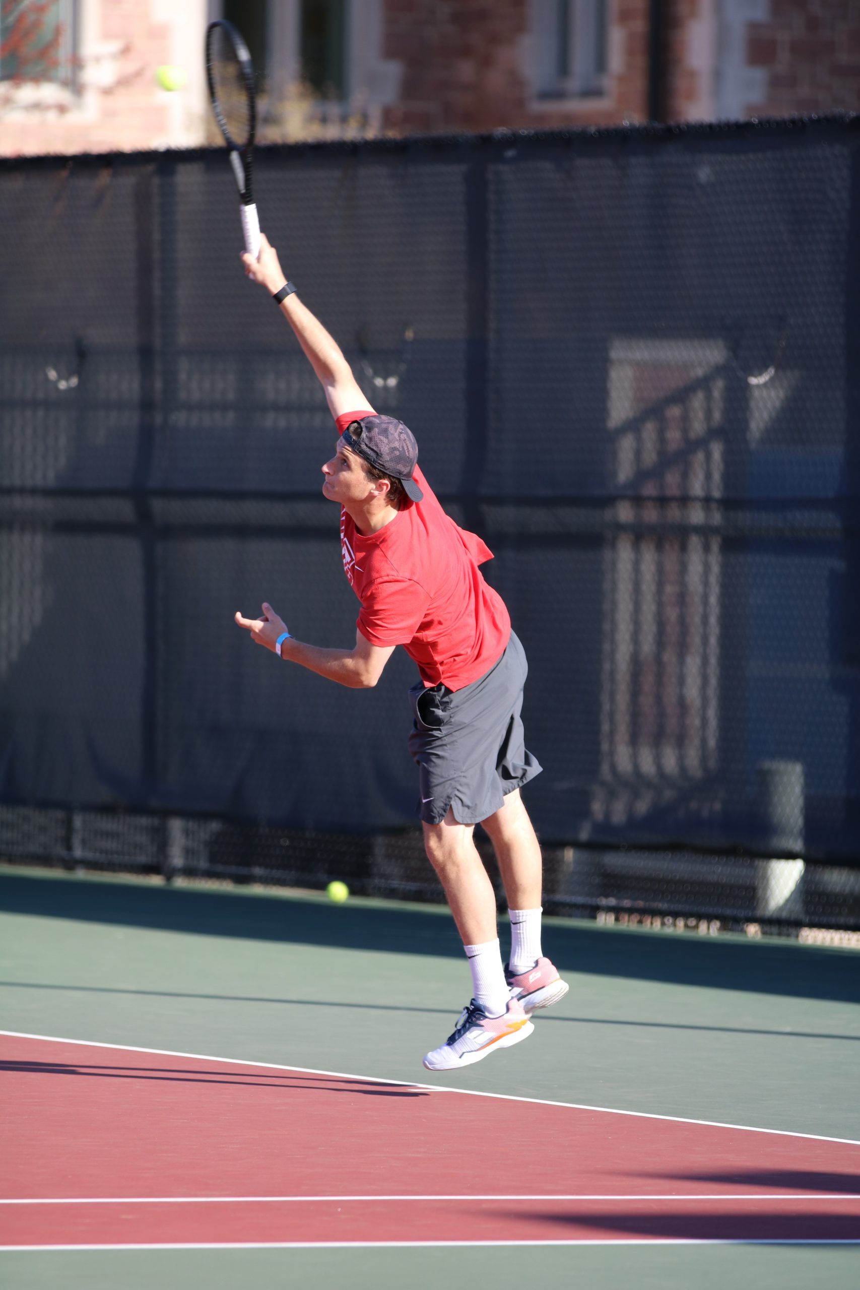 A player in a red shirt jumps with racquet held high to hit a tennis ball.