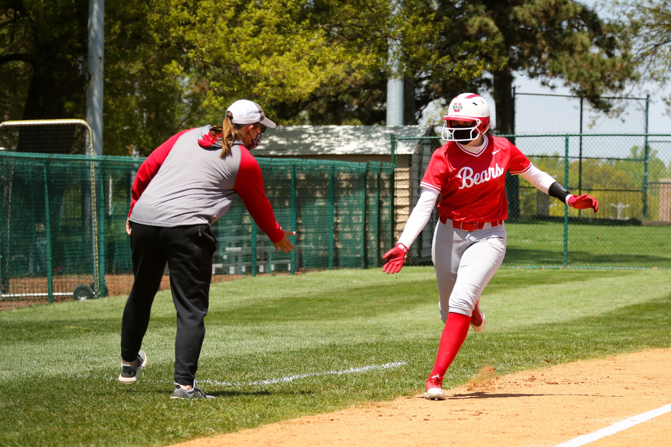 A softball player in a red jersey gives out a low-five to a coach in a red-and-gray shirt as she rounds the third base bag.
