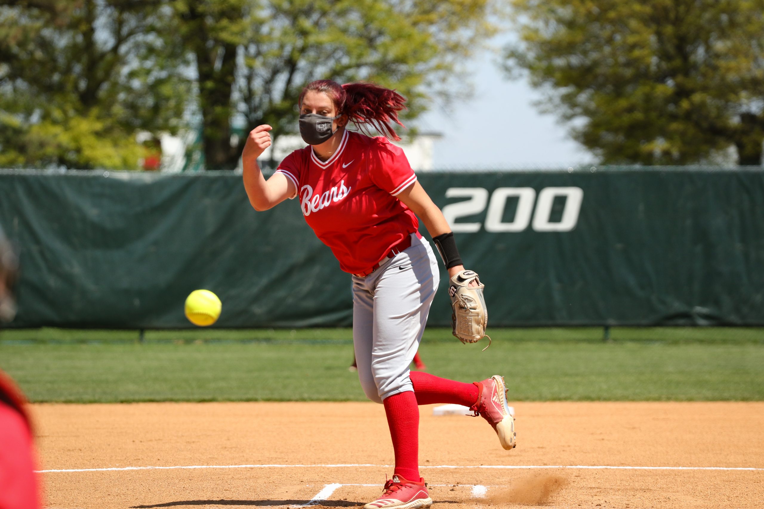 A softball pitcher in a red uniform and high red socks releases a softball toward home plate.