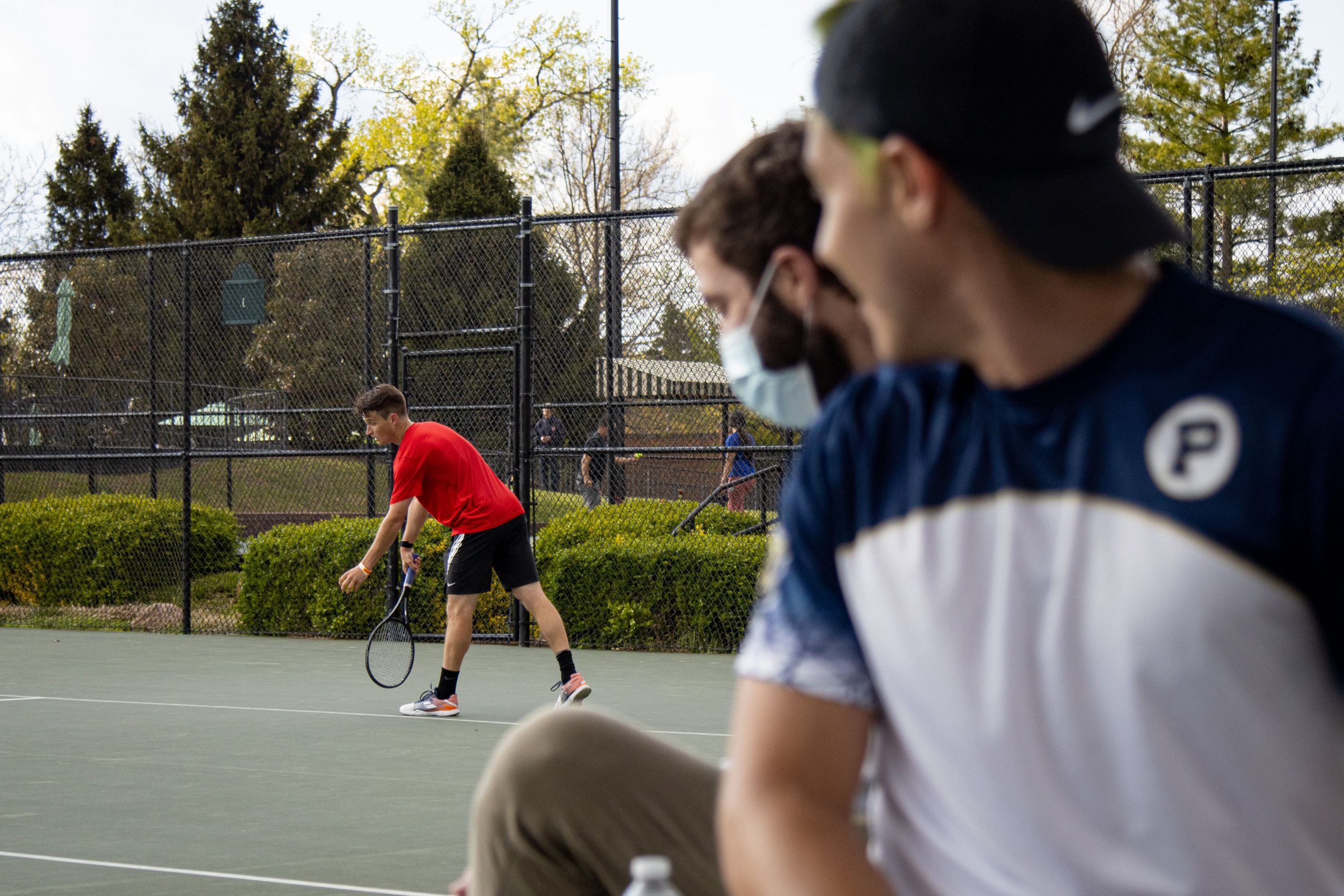 Two tennis players in white shirts look over their right shoulder toward a player in red who is bent over, preparing to serve.