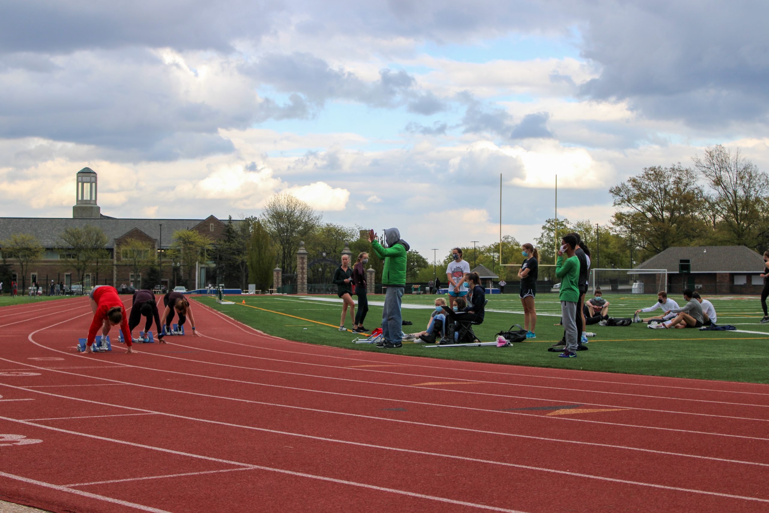 Three runners lean forward with their feet against blocks on a red track, as observers watch them from the green interior of the track underneath a partly cloudy sky.