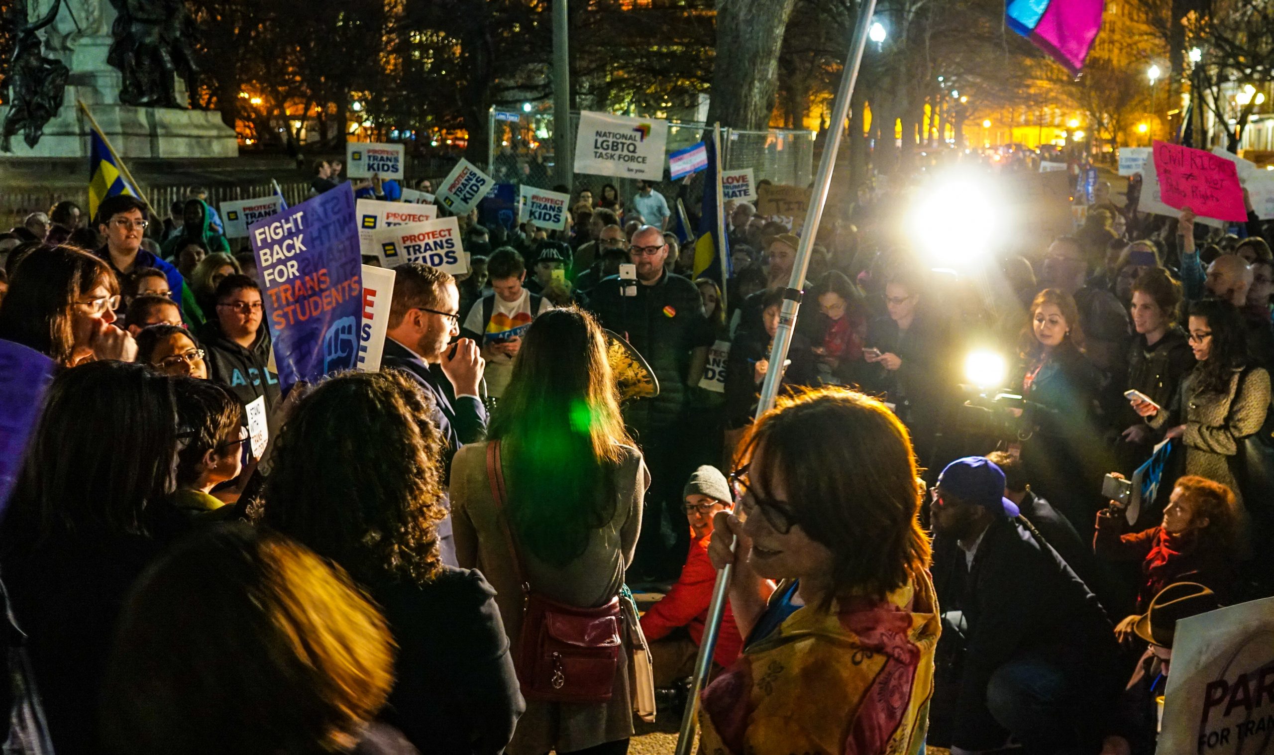 A group of protestors gather underneath a tree near a statue. People hold signs that say "Fight back for trans students" and "protect trans kids."