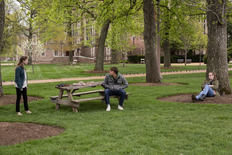 A man sits at a picnic table under trees, arguing with a woman standing on his left while another woman looks on from the right.