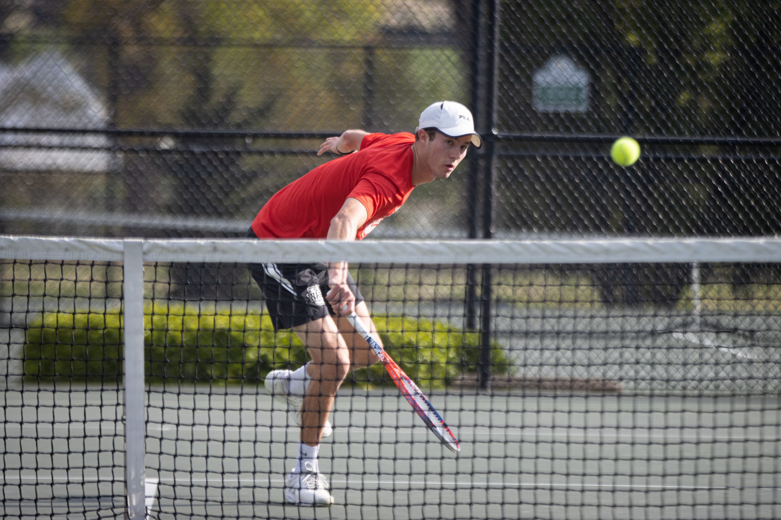 A tennis player in a red shirt lunges toward the net to corral a tennis ball.