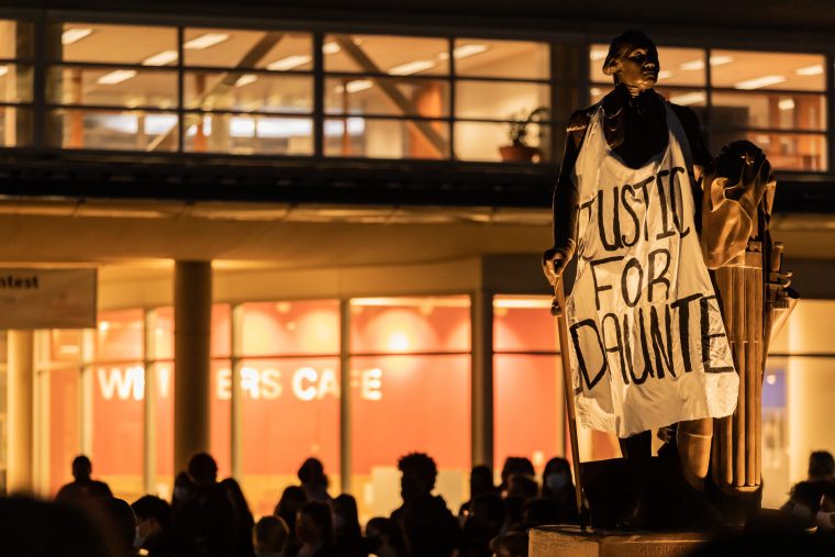 A white banner with the words "Justice for Daunte" is draped over a statue of George Washington, which stands in front of a glass paneled building. Silhouettes of a crowd of masked individuals can be seen near the statue.