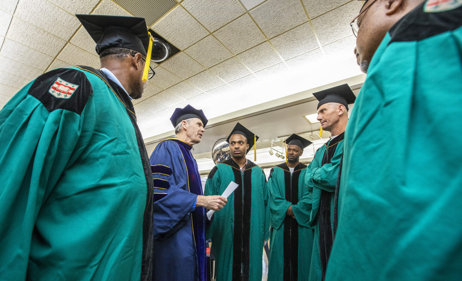 A man in blue academic robes speaks to a group of men in green robes and black graduation caps