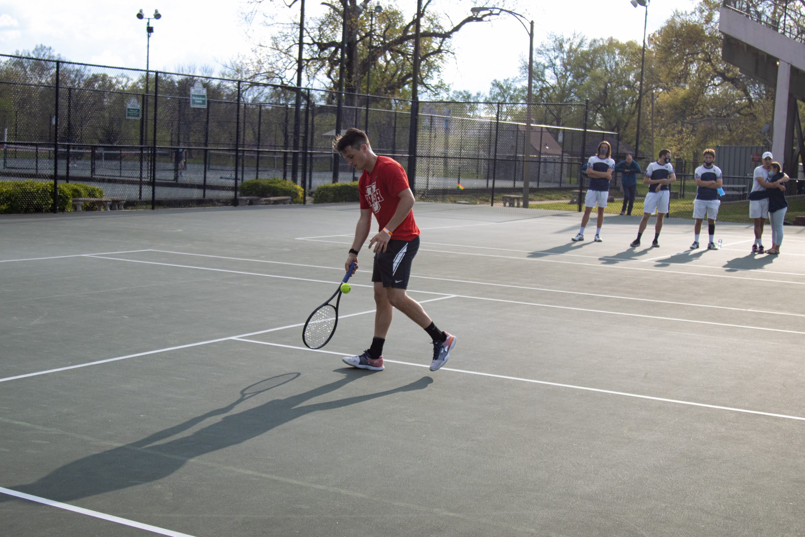 A tennis player wearing in red and shrouded in shadow holds his racquet in his right hand toward his outstretched right leg, as opponents in black uniforms watch from the upper right corner.