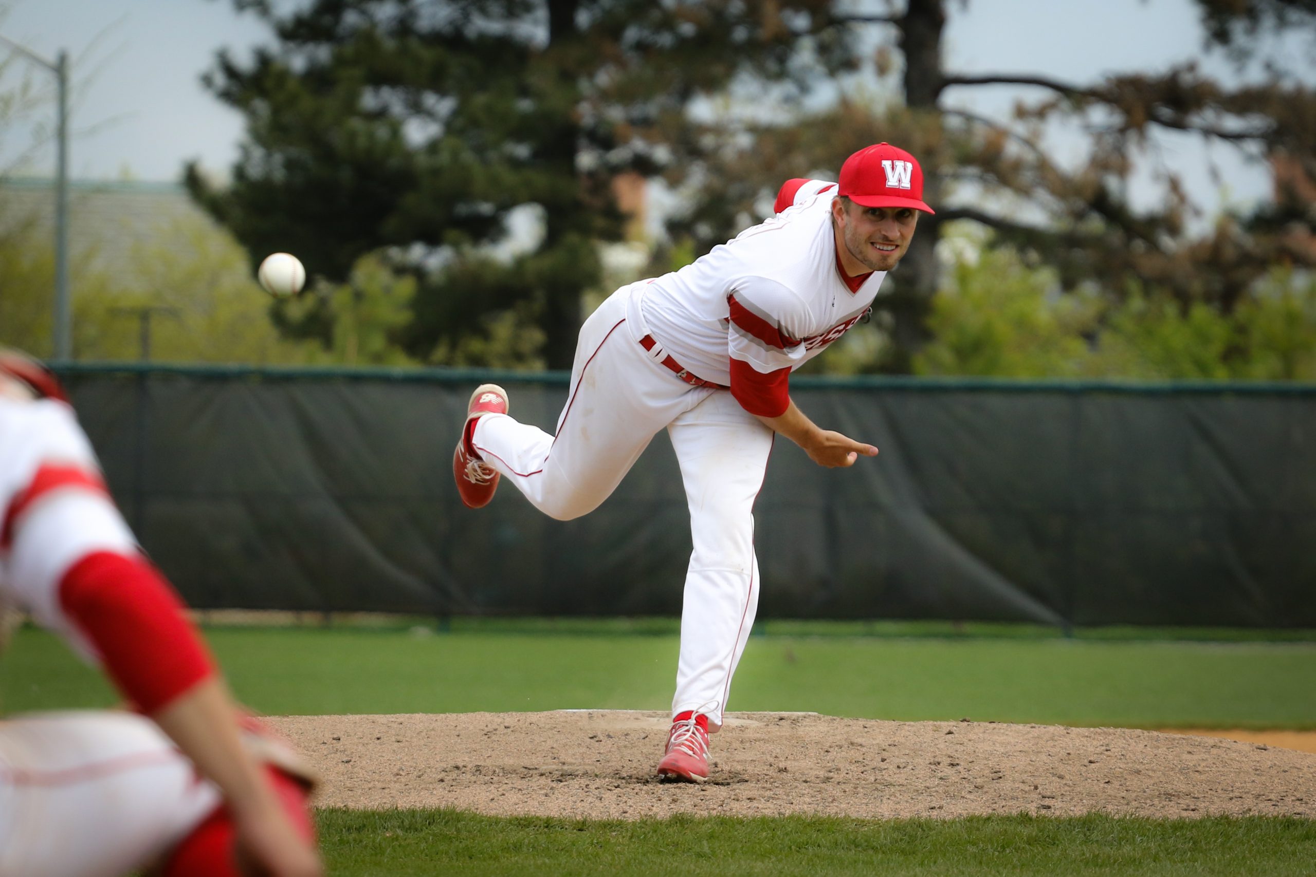 A pitcher in a white and red uniform grimaces after releasing a ball toward home plate. His right leg is in the air as he has just planted his left leg in front of him.