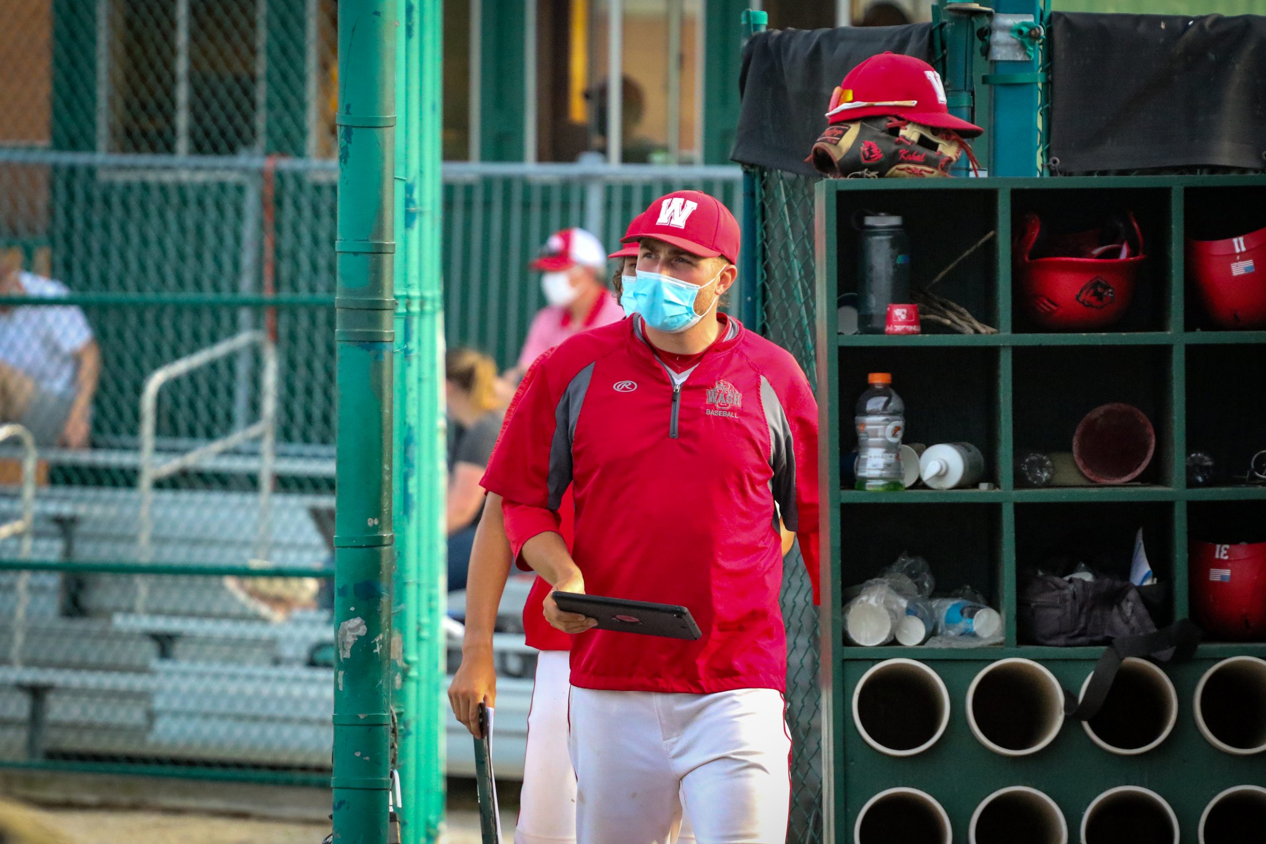 A player wearing a red jacket holds a tablet along the sidelines of a baseball field.