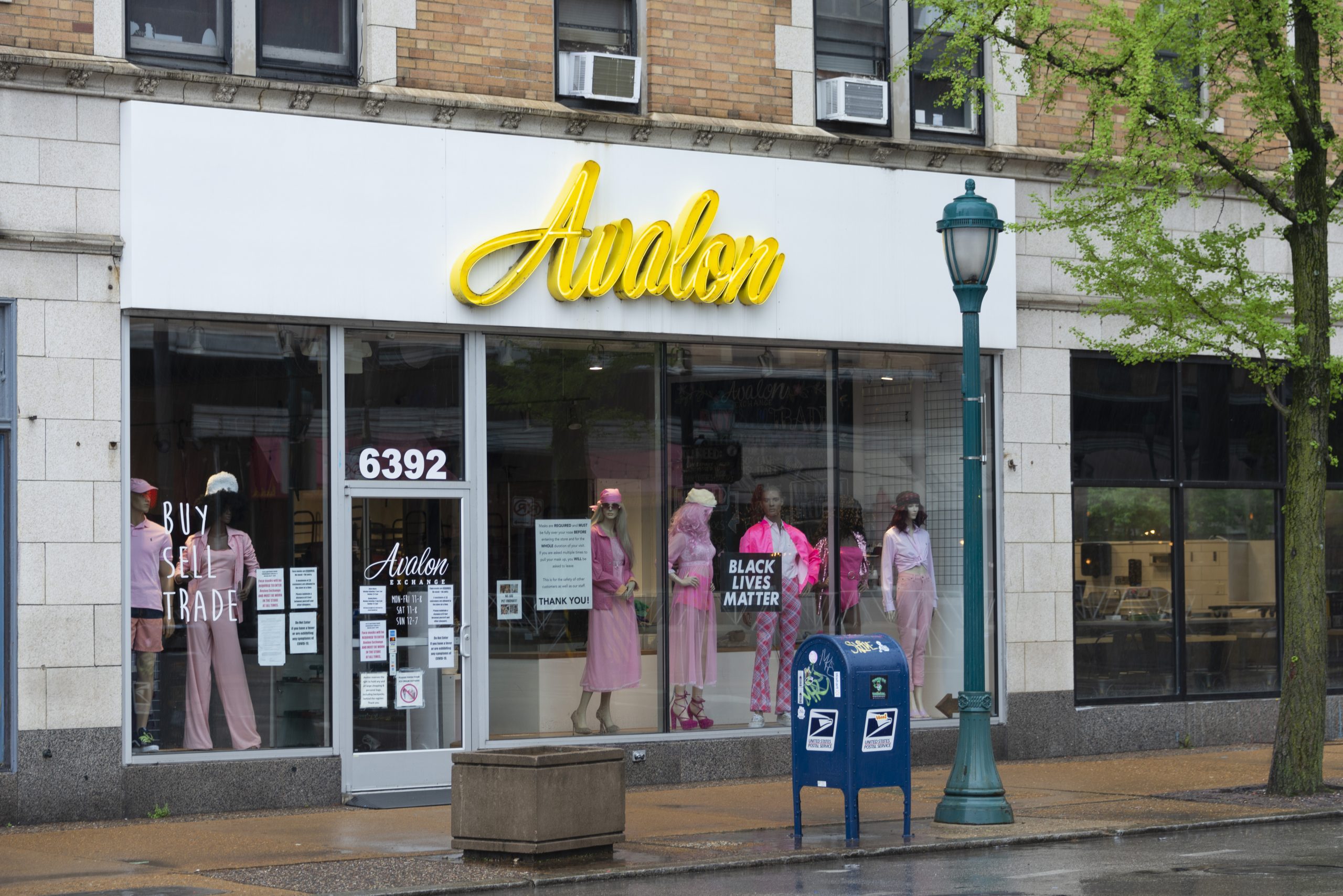 A storefront with floor-to ceiling glass windows stands behind a sidewalk. The word "Avalon" is written above the windows in large yellow cursive letters. Behind the windows, several mannequins are dressed in bright pink clothing. A black lives matter sign is also displayed behind a window.