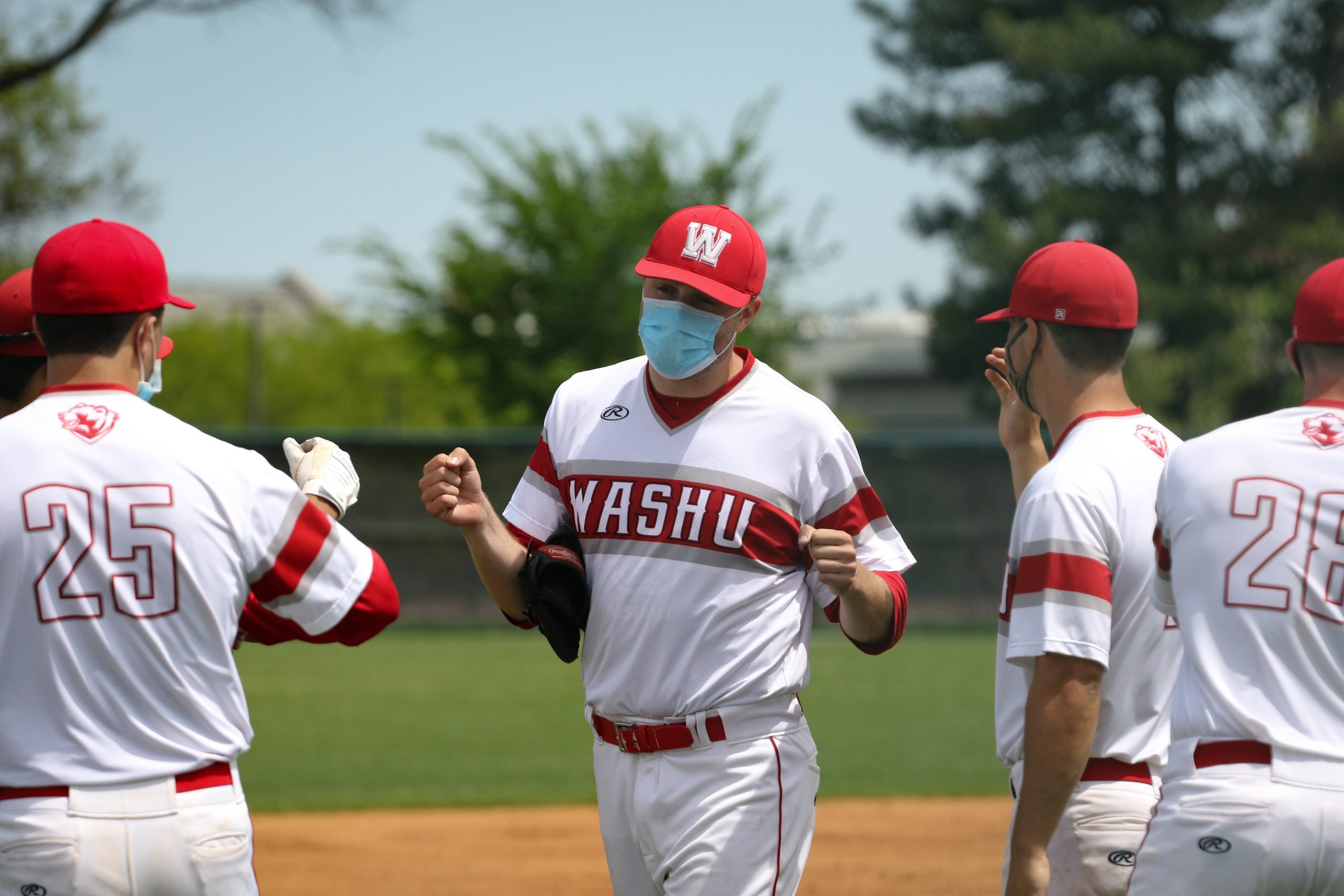 A player wearing a white and red uniform with a red cap, blue face mask and "WashU" written across the front of the shirt fist bumps two other players.