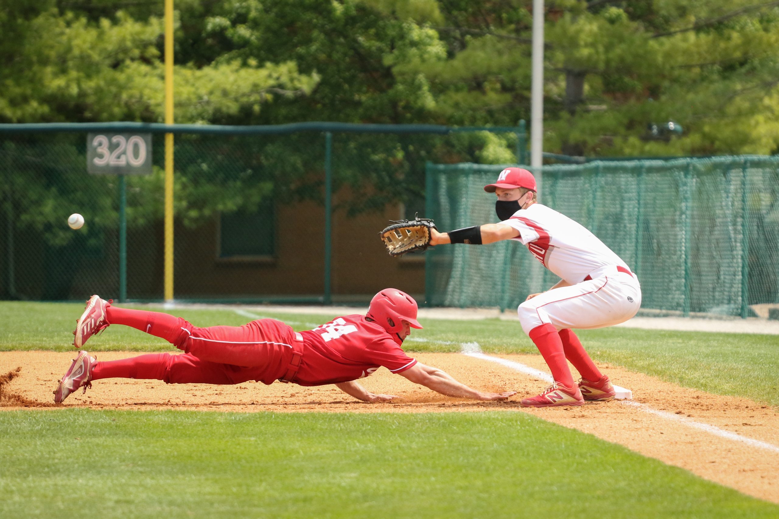 A player in a white uniform with his right foot on first base extends his left arm to catch a baseball as a player in a bright red uniform dives headfirst into the bag.