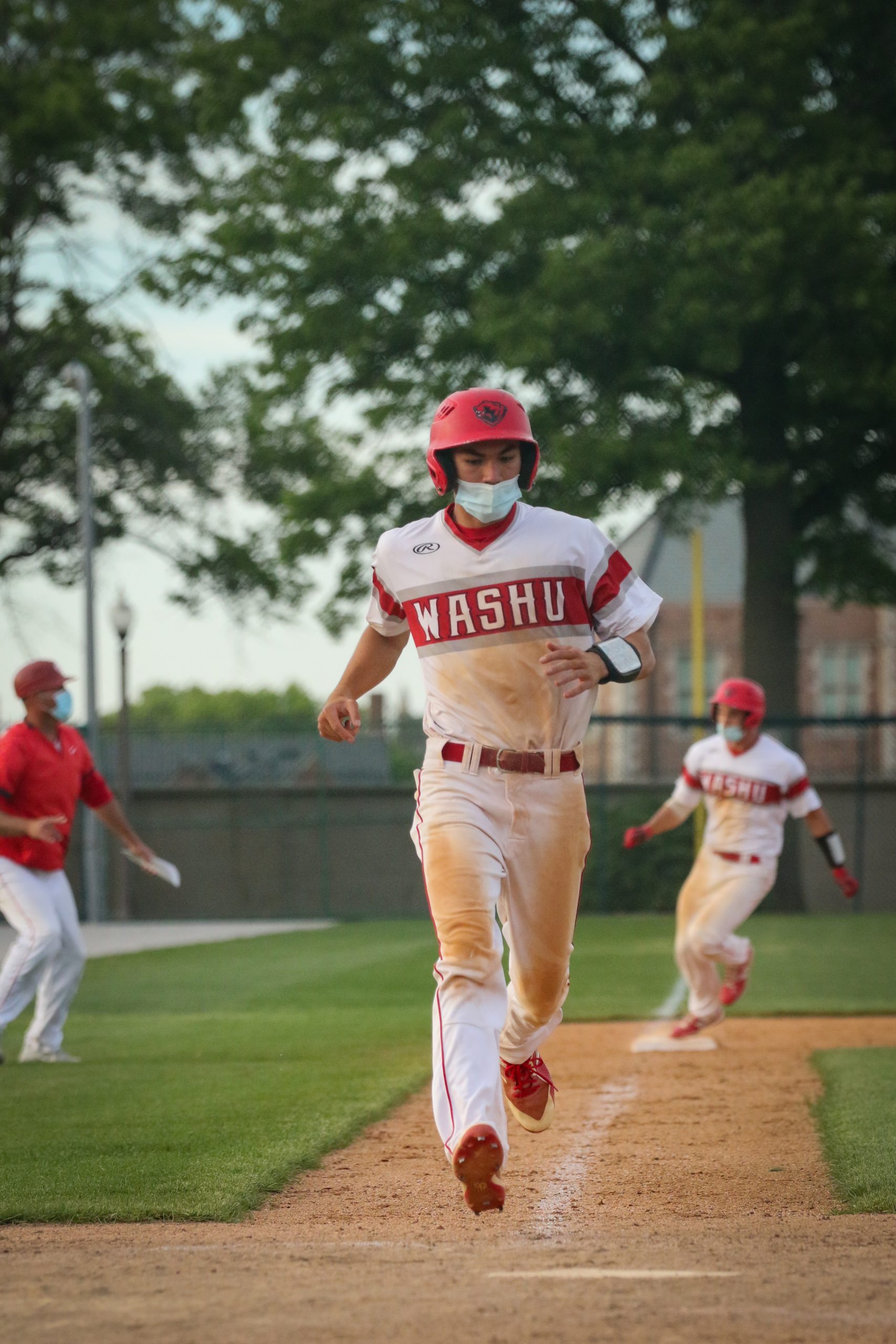 A player in a red and white uniform that reads "WashU" runs down the third base line as another player rounds third base behind him and a coach in a red shirt looks on from the left side.