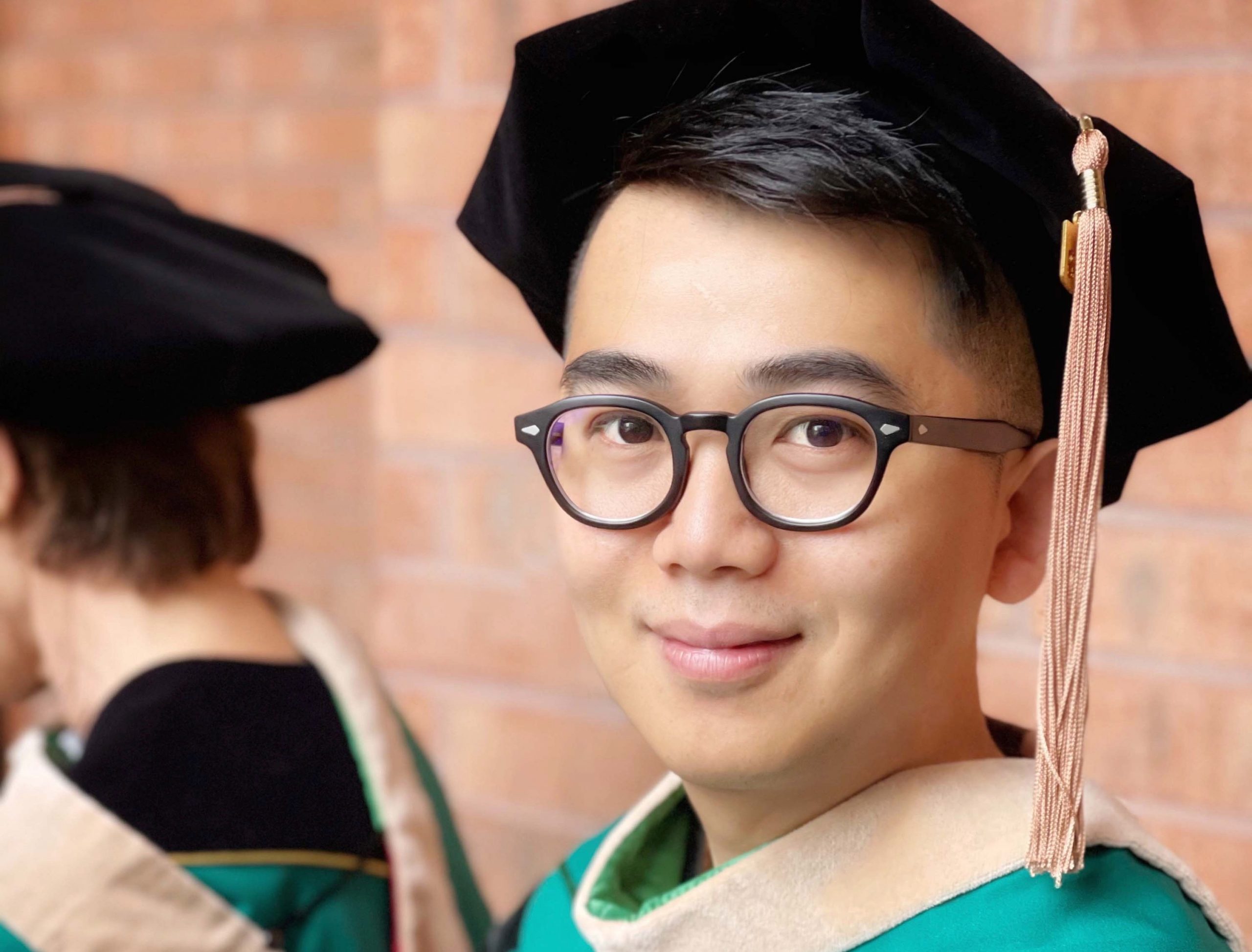 A smiling man wears a black graduation cap and green gown.
