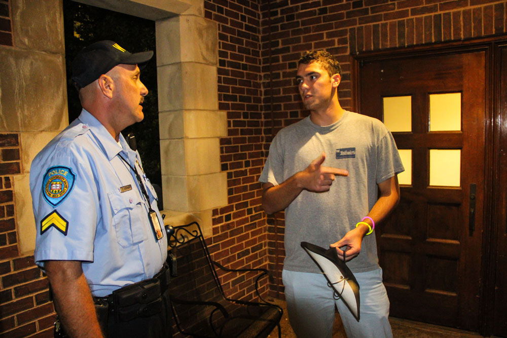 A police officer in a light blue shirt and black cap faces a student wearing a gray shirt. The two figures stand in front of a brick building.