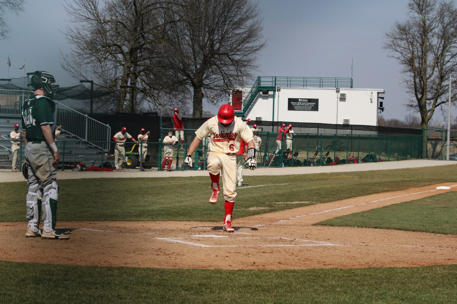 A player in a white uniform and red helmet raises his right foot to stomp on home plate near a dirt infield in front of blue sky.