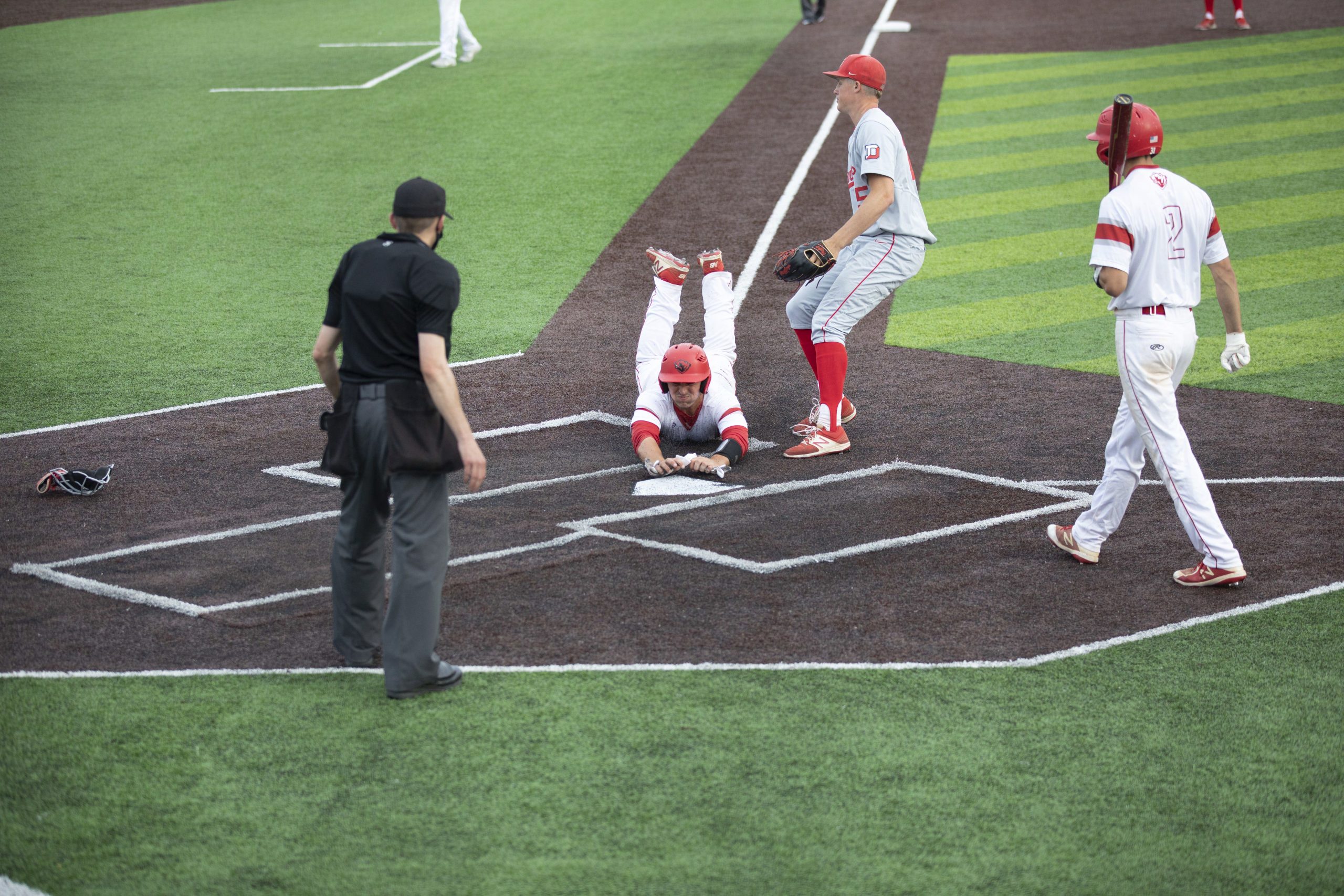 A baseball player in a white uniform and red helmet dives into home plate as an opponent in a gray uniform looks on from the right side and an umpire in black watches from the left.