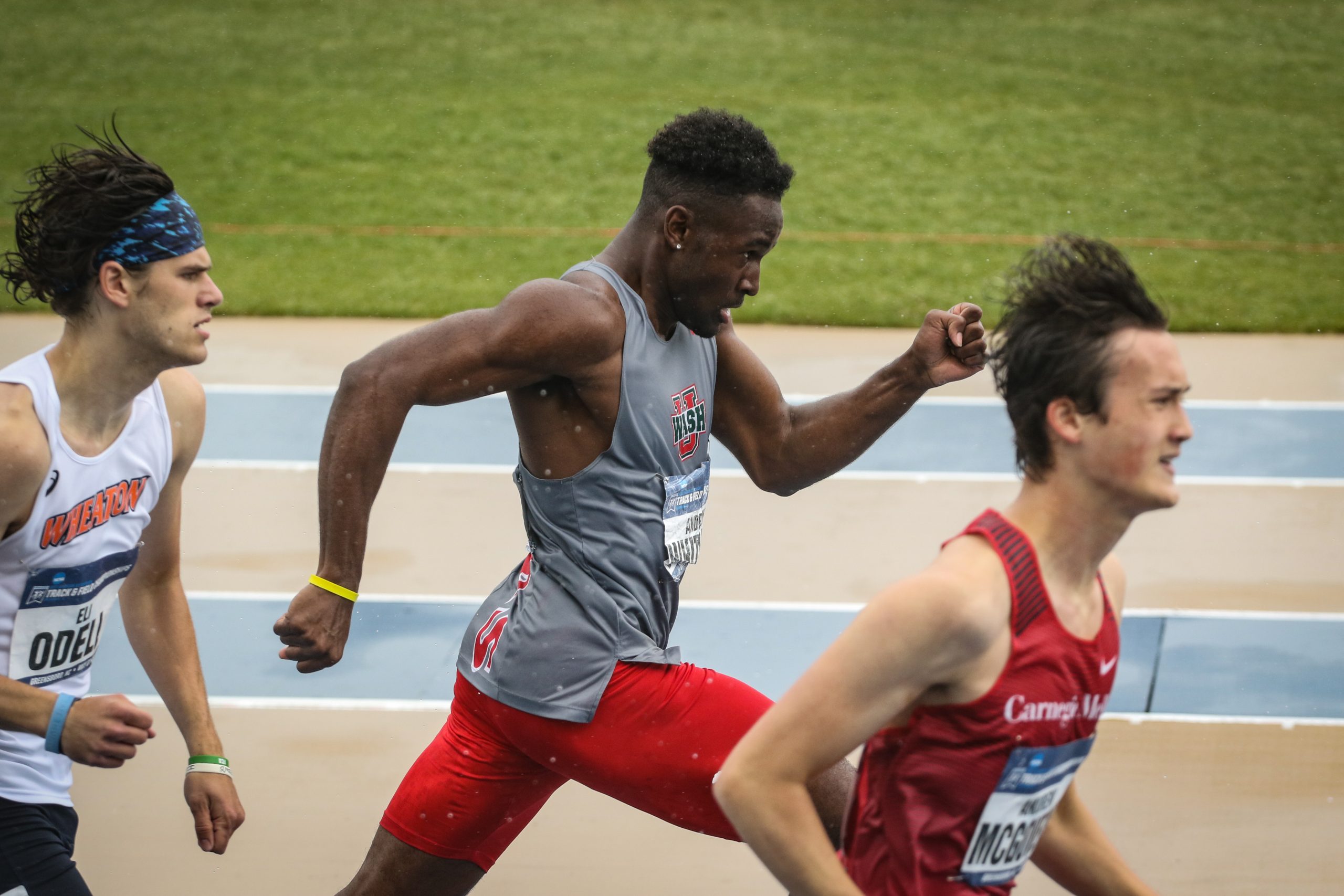 A runner wearing a gray tank top and red shorts strides forward between two other runners, with his left arm forward and his right arm back on a beige and blue track with grass in the background.