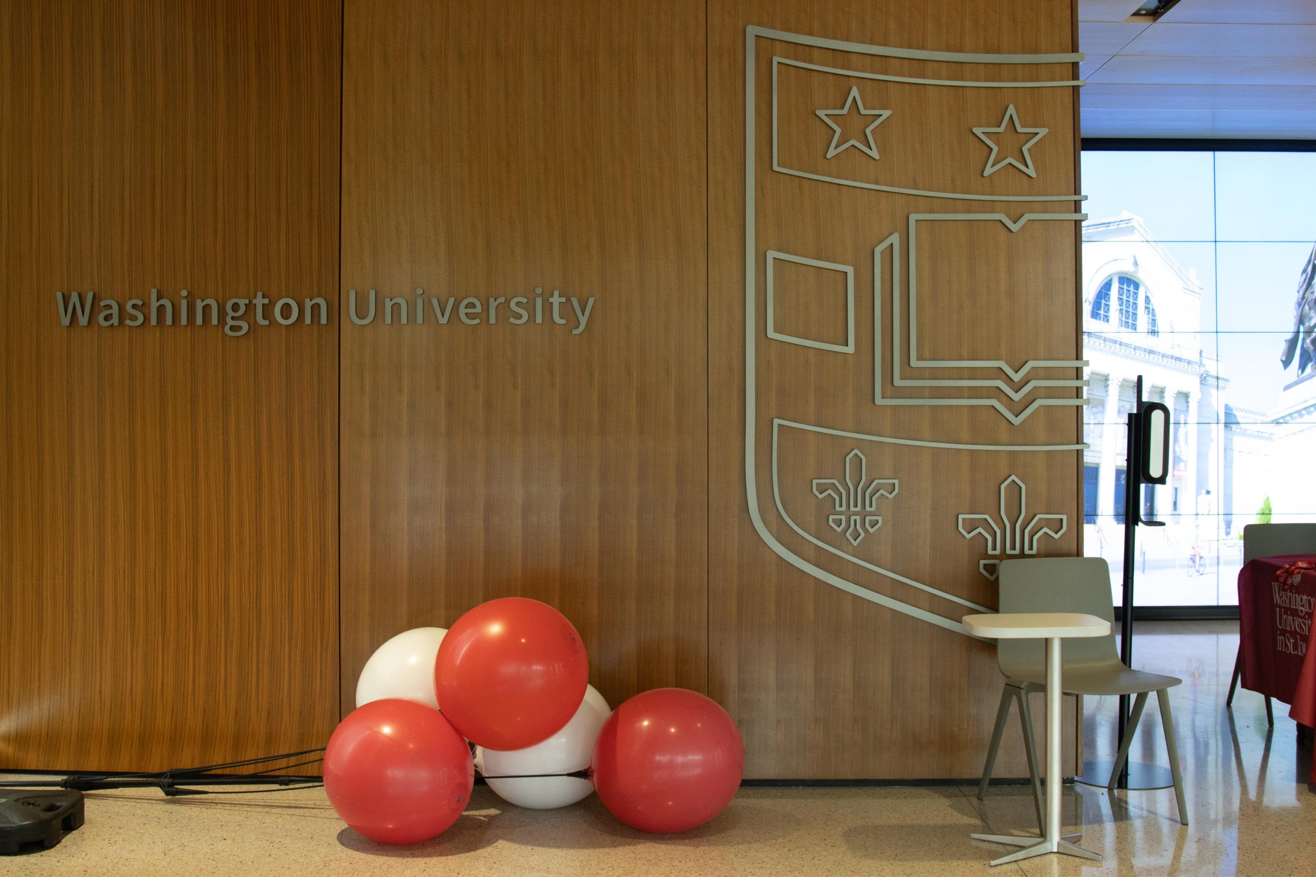 Red and white balloons sit on the floor in front of a wood-paneled wall labeled "Washington University" with the university's seal.