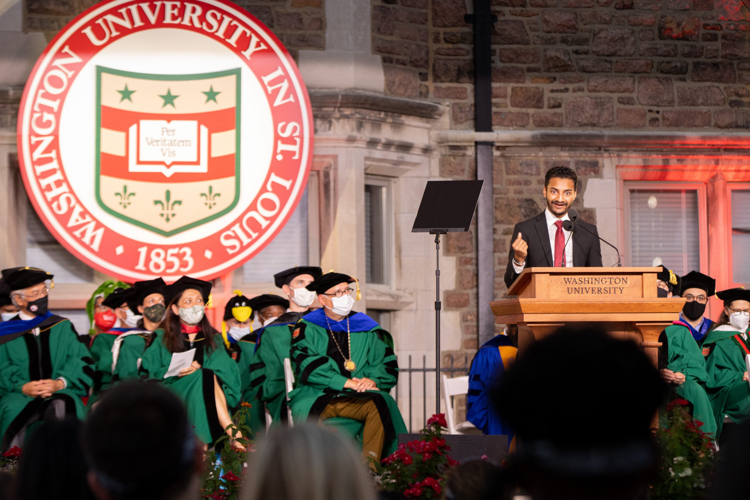 A man in a dark suit with a red tie stands at a wooden podium with "Washington University" written on it. To his left, people in green robes, black caps and masks sit in folding chairs. The Washington University seal hangs above their heads.