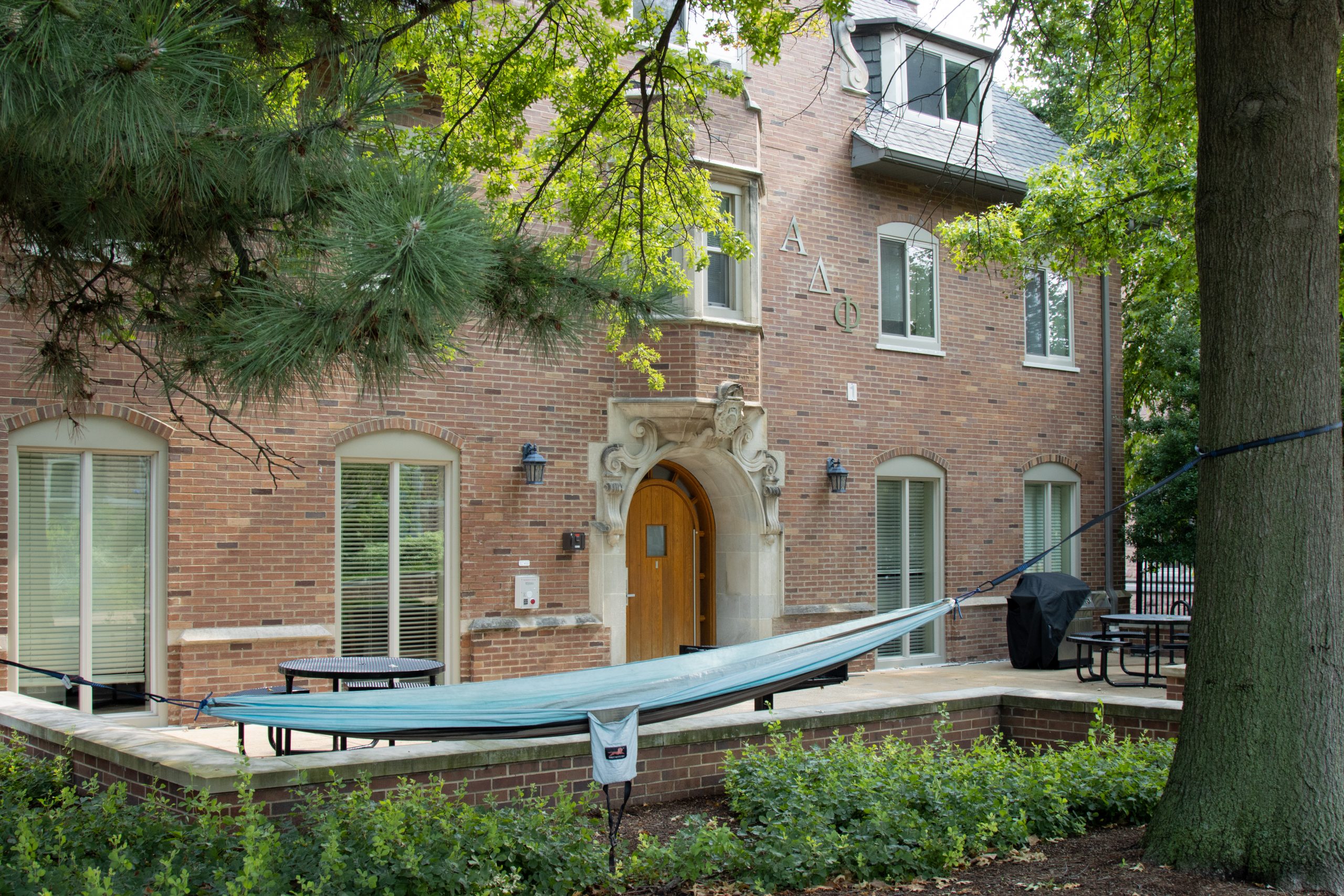 A green hammock hangs between two trees in front of a brick building with a brown wooden door. Greek letters can be seen above the door.