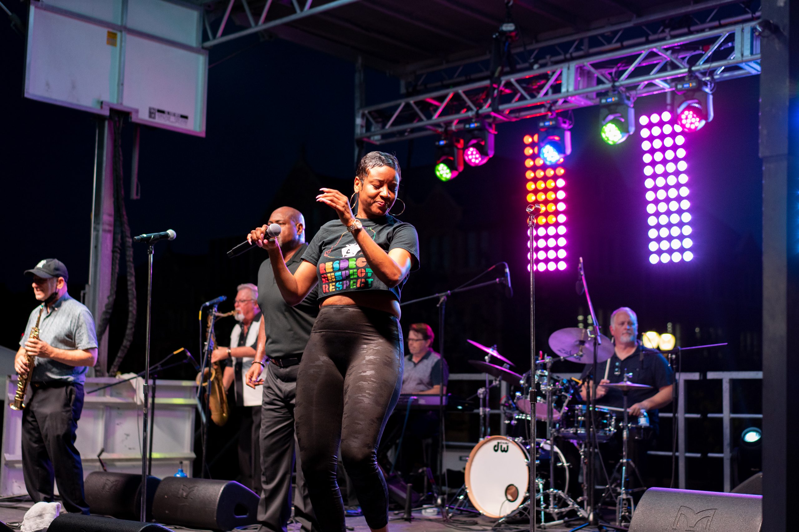 A woman in leggings and a black shirt reading "respect" stands on a stage holding a microphone. Behind her, men in dark clothes play horns and drums, while purple lights illuminate the stage.