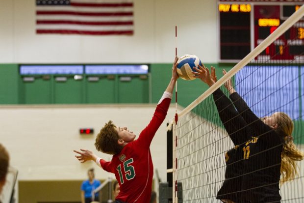 A volleyball player in a dark red jersey extends their right arm toward the net to block a volleyball that is being hit downward by a player on the other side of the net wearing a black uniform. An American flag hangs on the wall in the background.