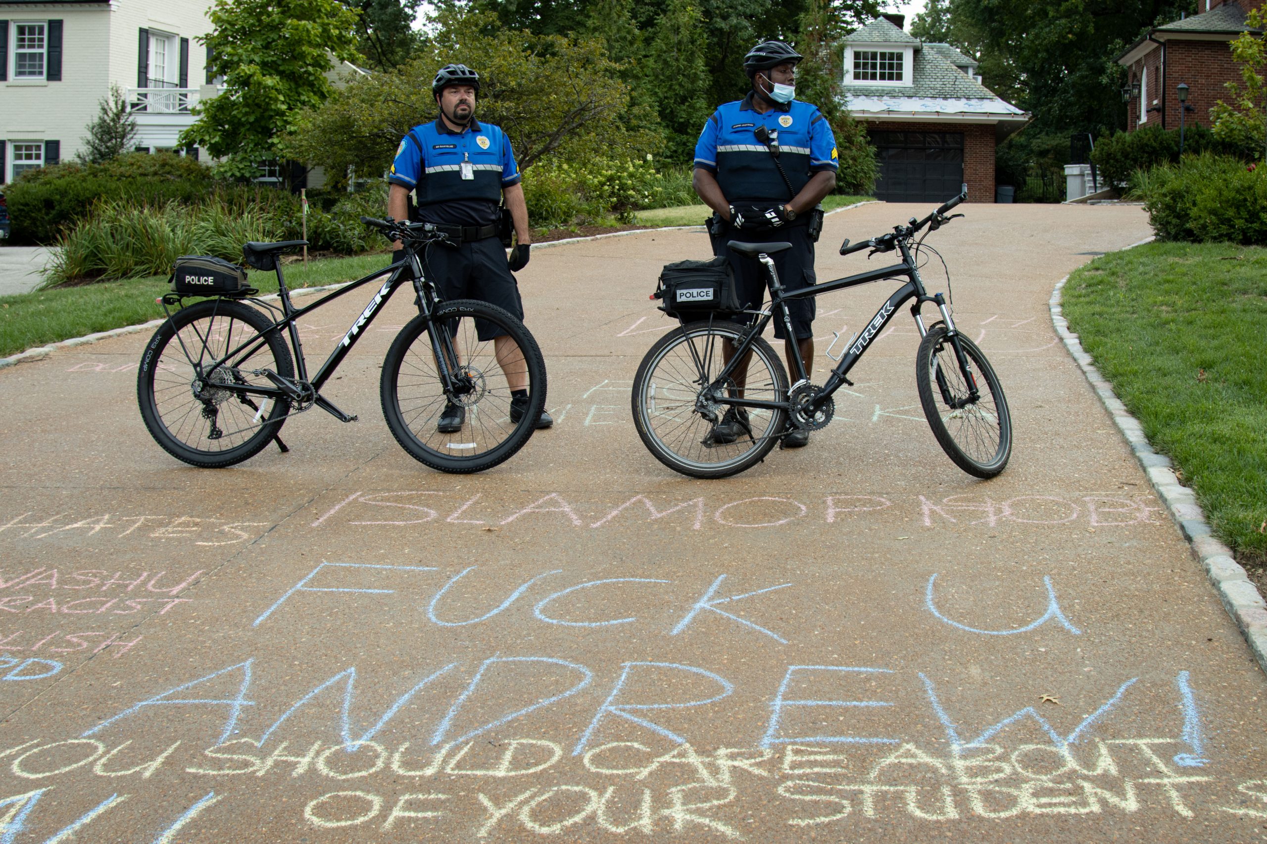 Two police officers in dark blue shirts and black shorts stand behind bicycles on a brick driveway. Written in chalk on the driveway in front of them are slogans such as "Fuck u Andrew!" "Islamophobe," and "You should care about ALL of your students." There are trees in the background.