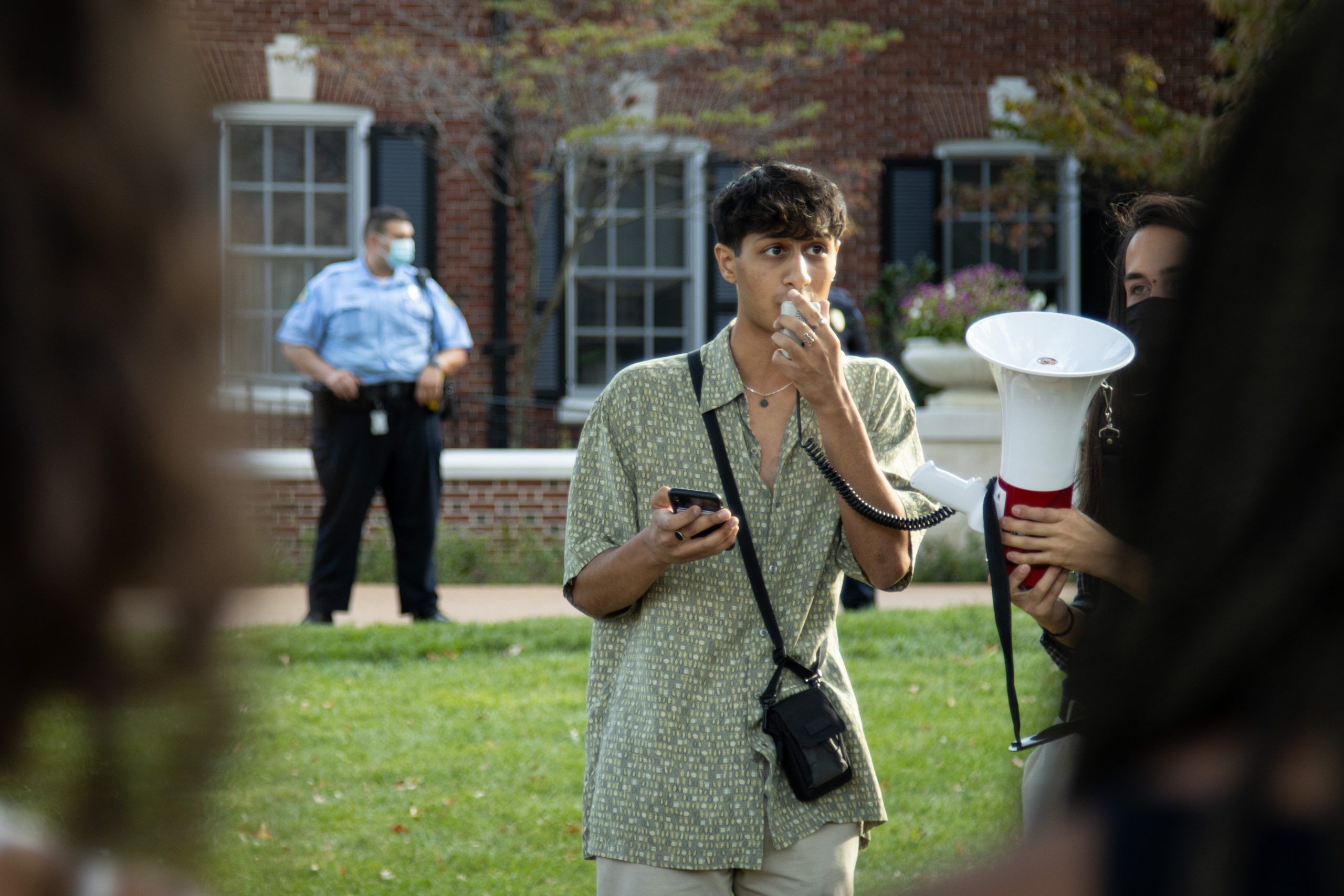 A student wearing a patterned green button shirt stands with a microphone in his left hand held up to his mouth, a phone in his right hand held near his chest and a black camera draped around his neck. A police officer in light blue shirt and black pants stands on the left in front of a brick building in the left of the background.