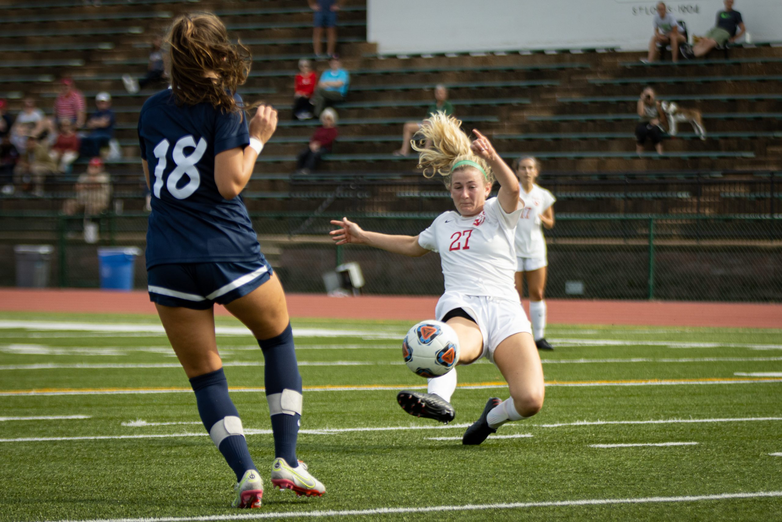 A soccer player in a white jersey extends her right foot to kick a white and red soccer ball. Her left foot curls behind her as she nearly parallels the ground and her blond hair flows above her head. A player in a blue uniform stands on the left side, moving to the left.