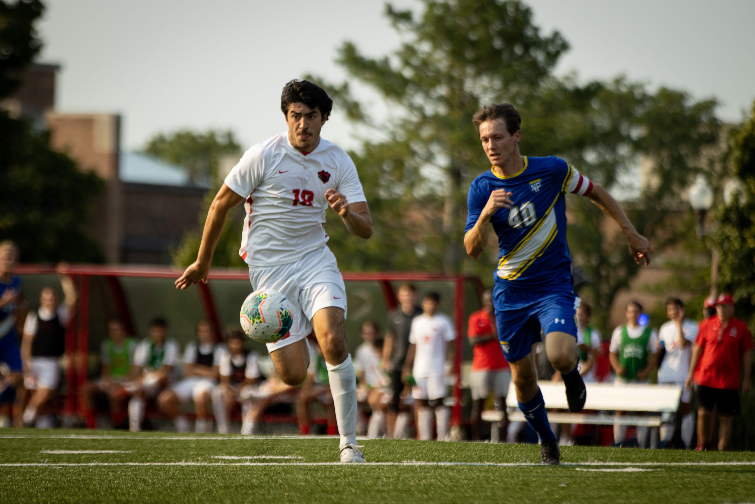 A soccer player wearing a white jersey with a red number 18 chases after a soccer ball as a player in a blue uniform with a gold stripe chases after him.