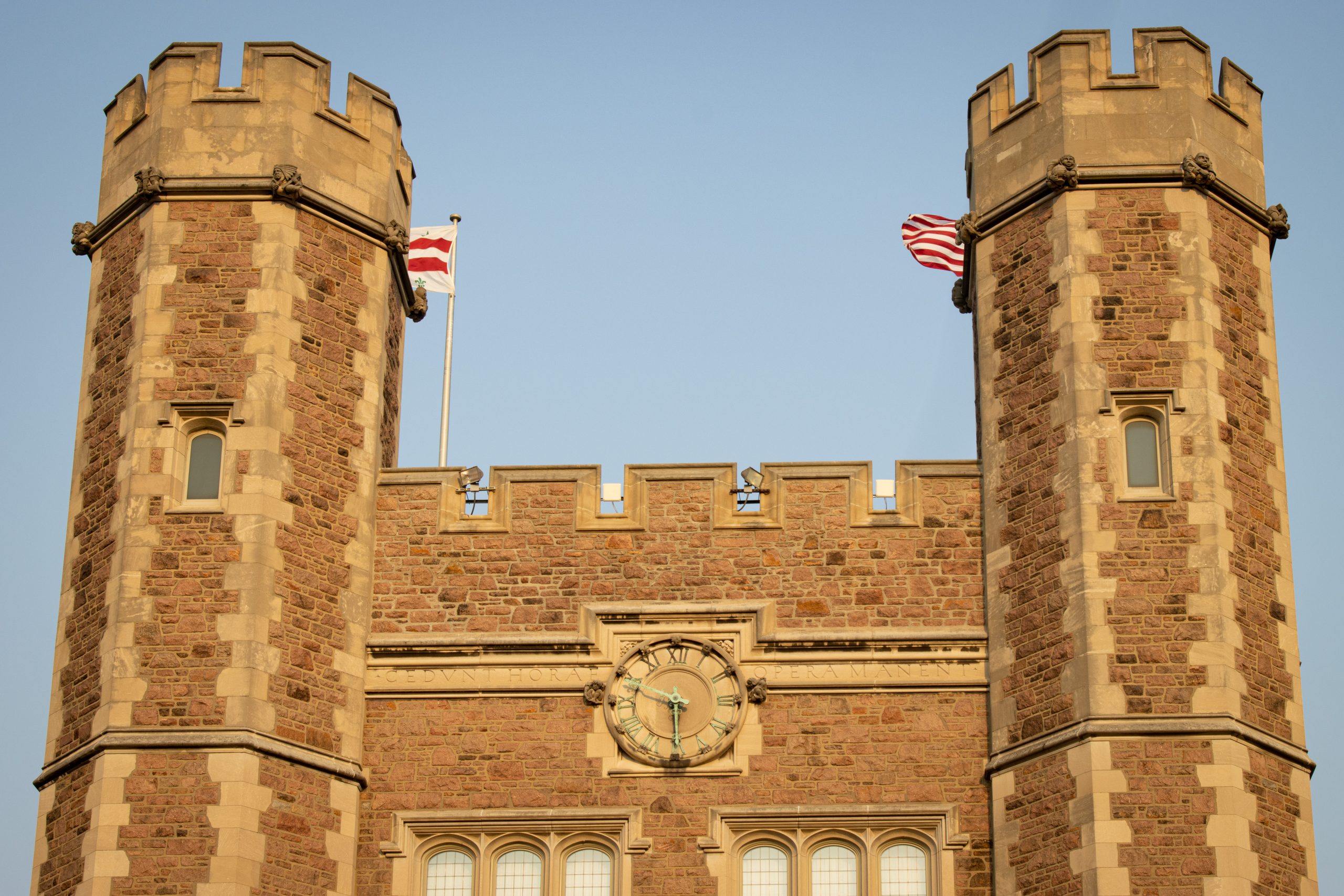 A red brick facade of a building with two round towers stands in front of a pale blue sky. A clockface and windows can be seen in the center of the building, while two red and white flags can be somewhat seen behind the towers on each side.