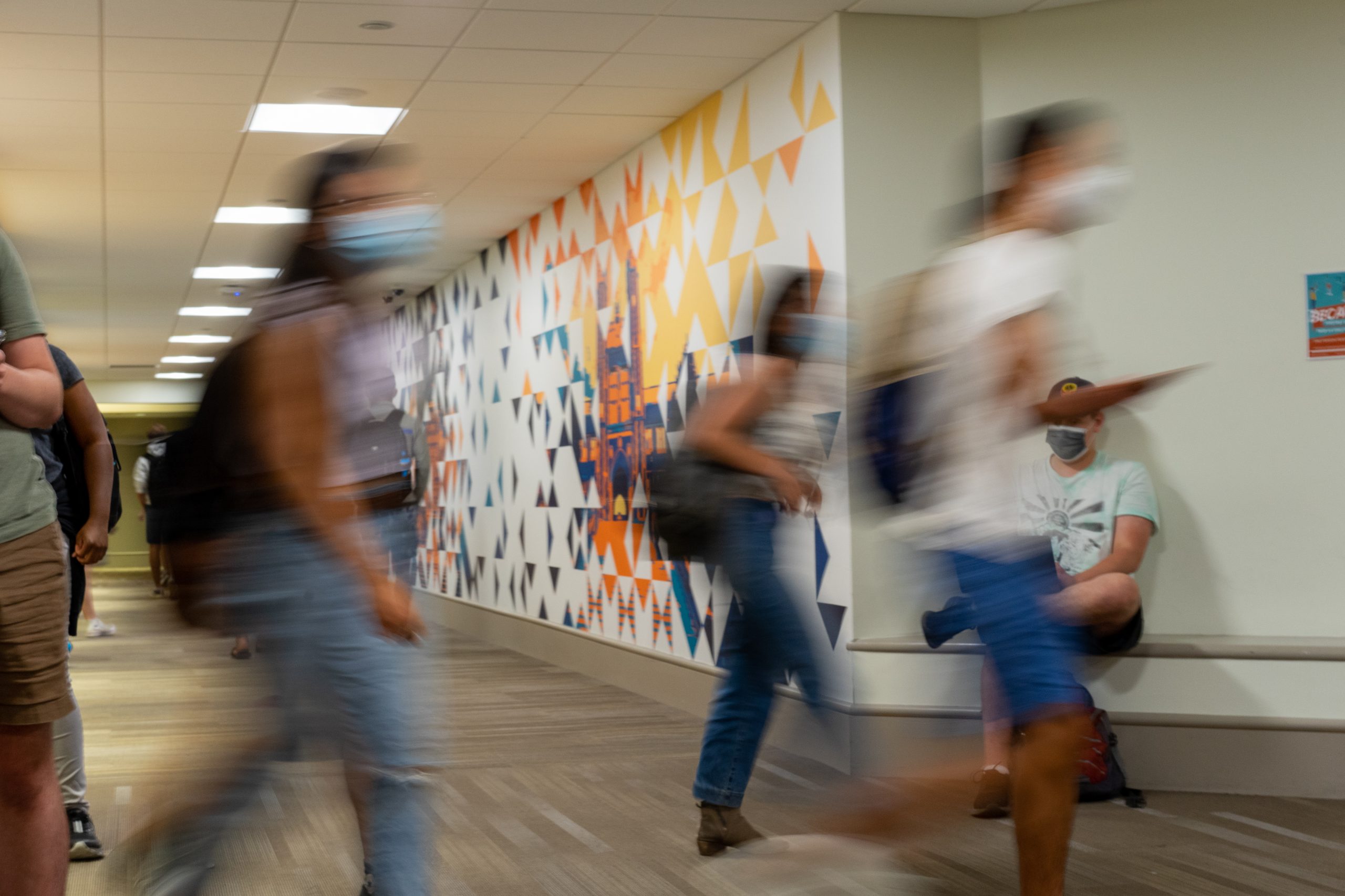blurry outlines of masked people walk across a hallway with a grey floor, green walls and lights on the ceiling.