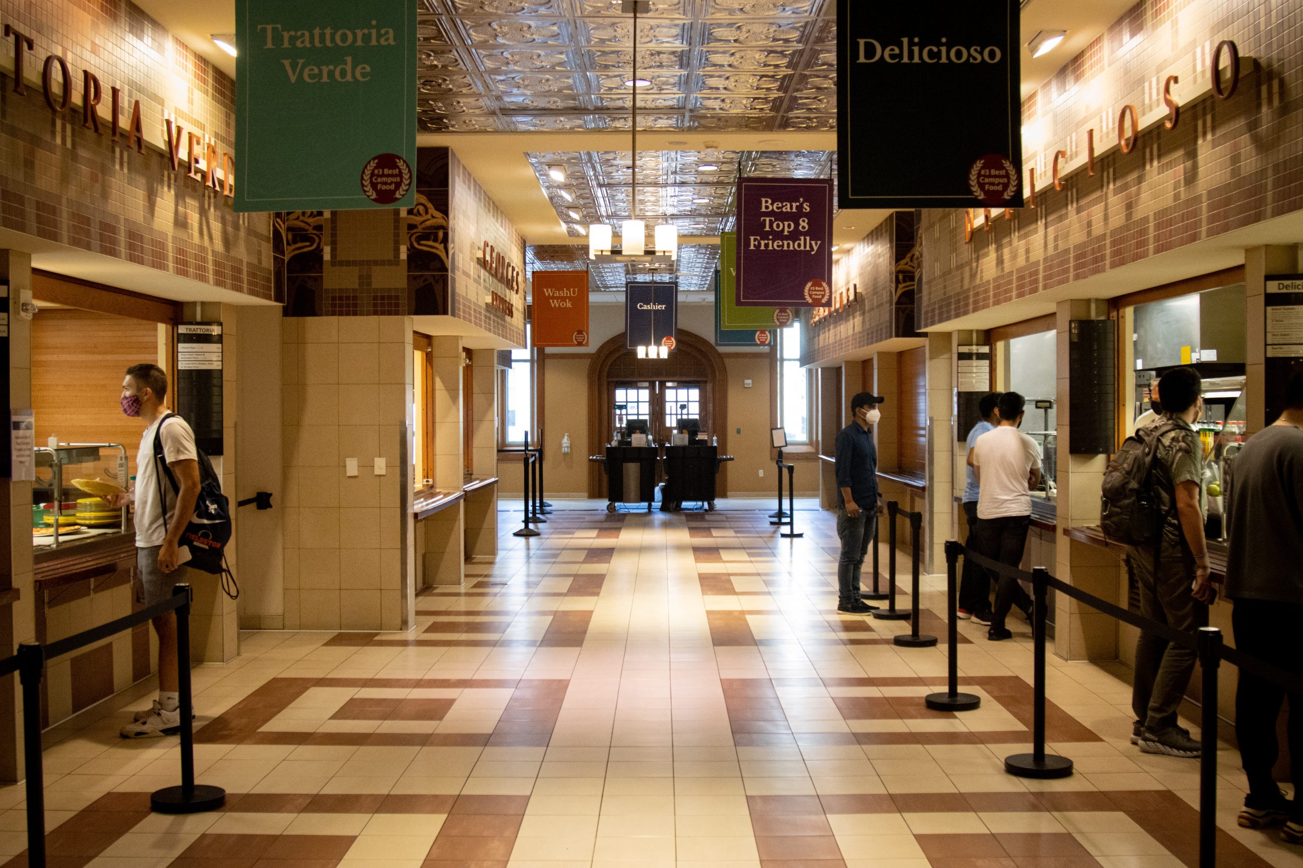 People wearing masks stand on either side of a hallway with a brown and beige tiled floor, facing windows with trays and food items in them. Banners reading "Trattoria Verde," "Delicioso" and "Bear's Top 8 Friendly" hang from the ceiling.