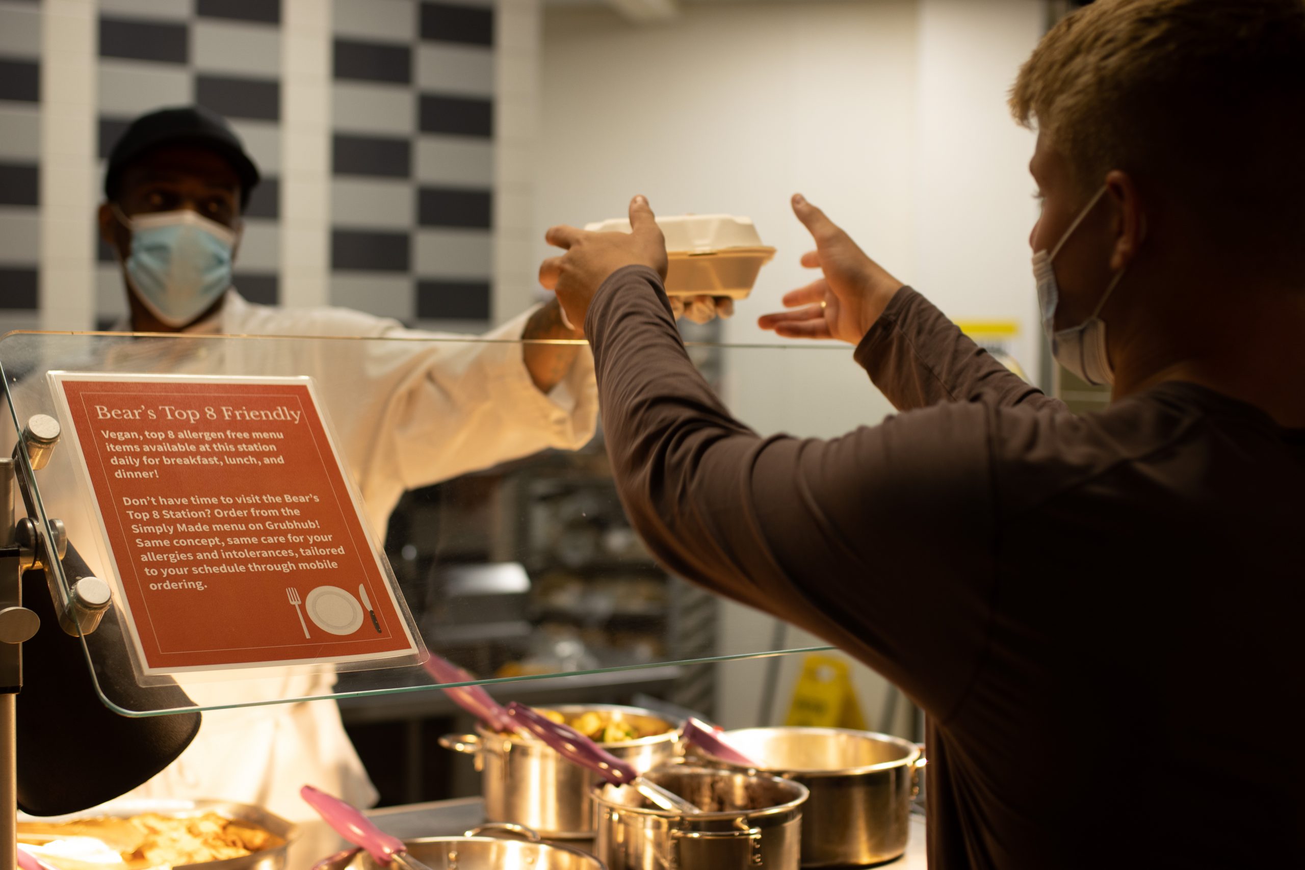 A man in a white coat hands a student a box of food near a sign reading “Bear’s Top 8 Friendly.”