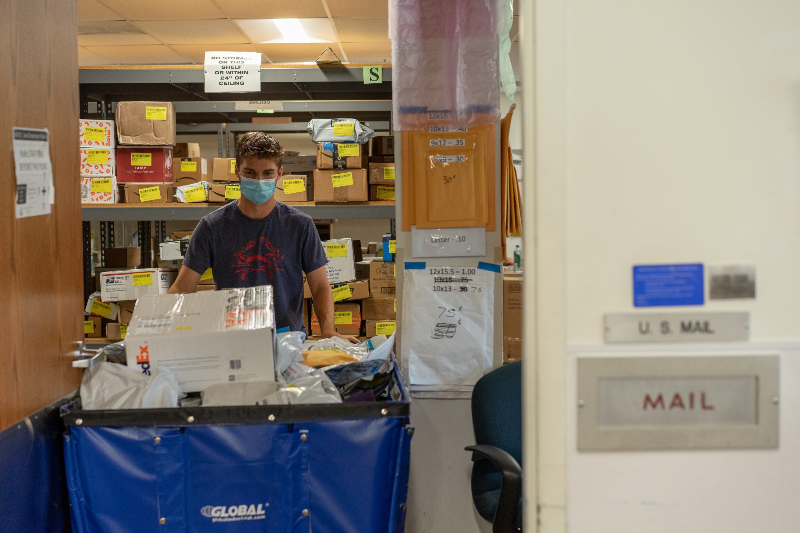 A student wearing a mask pushes a large cart full of cardboard packages.