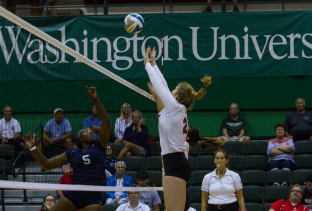 A volleyball player in a white shirt and black shorts jumps near the right side of a volleyball net, reaching up with both arms to hit a blue and white volleyball.