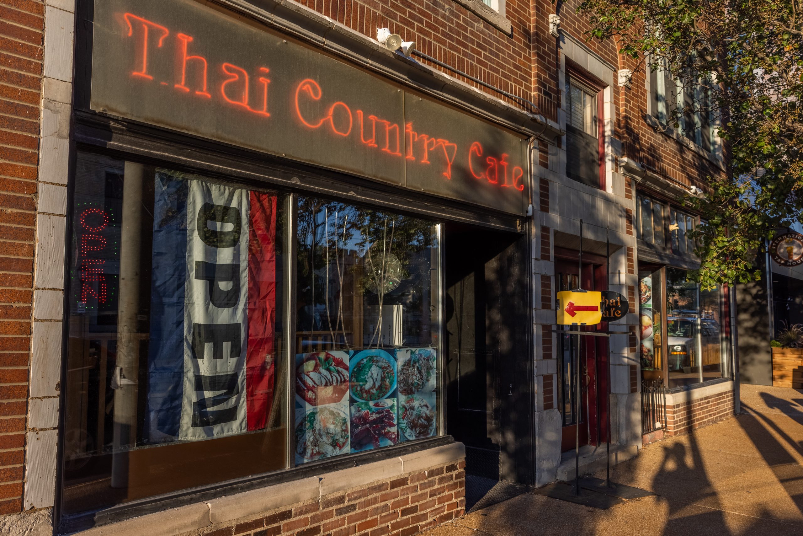A brick storefront displays a neon sign reading "Thai Country Cafe" and a flag reading "OPEN."
