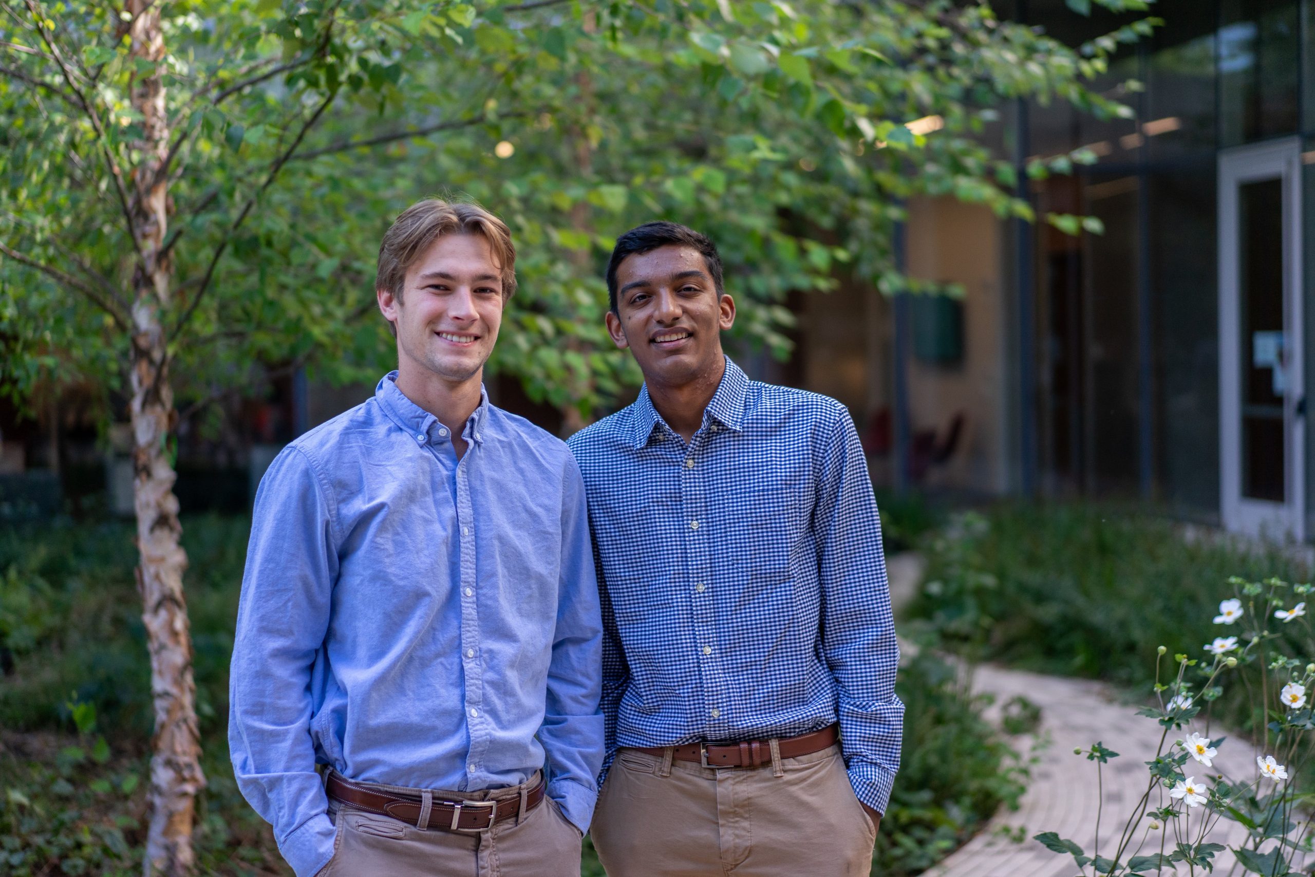 Two men wearing blue patterned button down shirts and khaki pants stand next to each other and smile. Trees, grass and a stone path can be seen in the background.