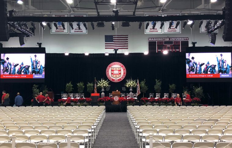 Rows of empty white folding chairs sit on a grey carpet, facing a stage with silver chairs, red roses, a wooden podium and a large seal with the WU logo. TV screens, lights and a curtain hang behind the stage.