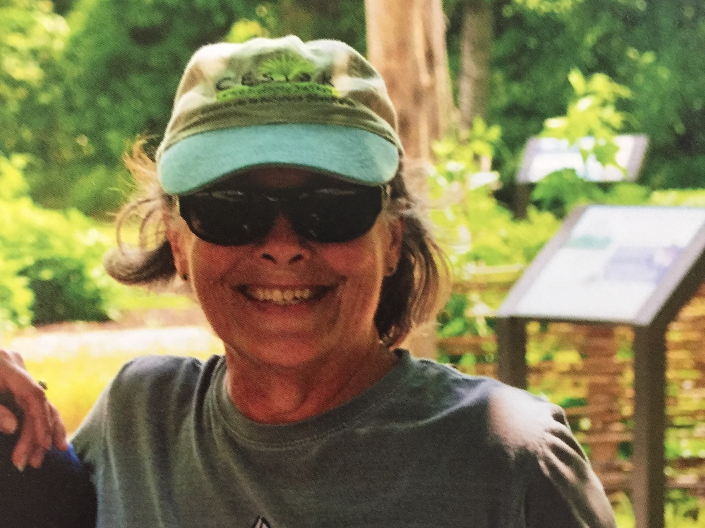 A woman in sunglasses and a green baseball cap smiles, surrounded by nature.