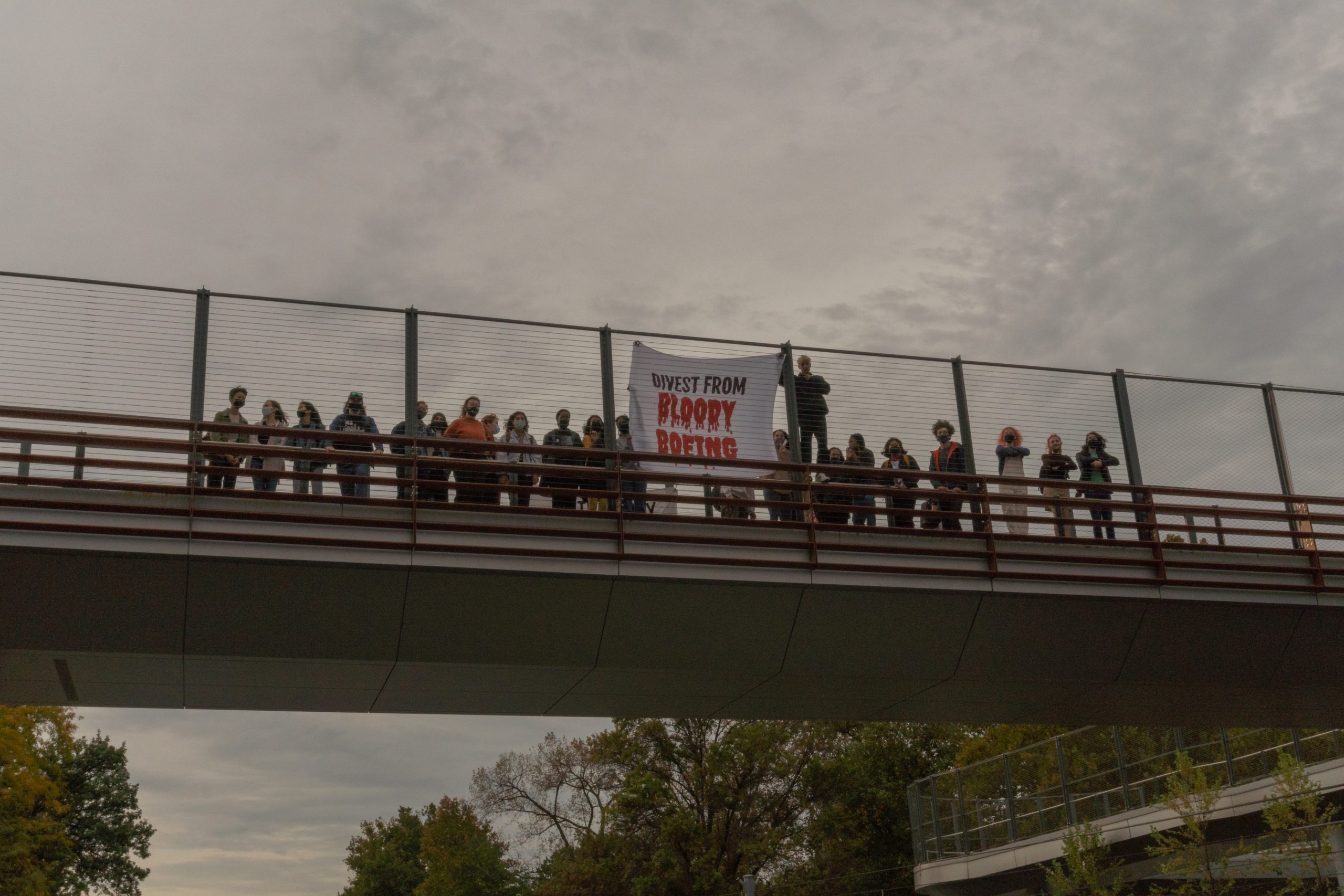 A crowd of people stand behind a chain-link fence on a walkway suspended in the air. A white sign with "divest from bloody Boeing" written in red with blood drops on the letters is held behind the fence.