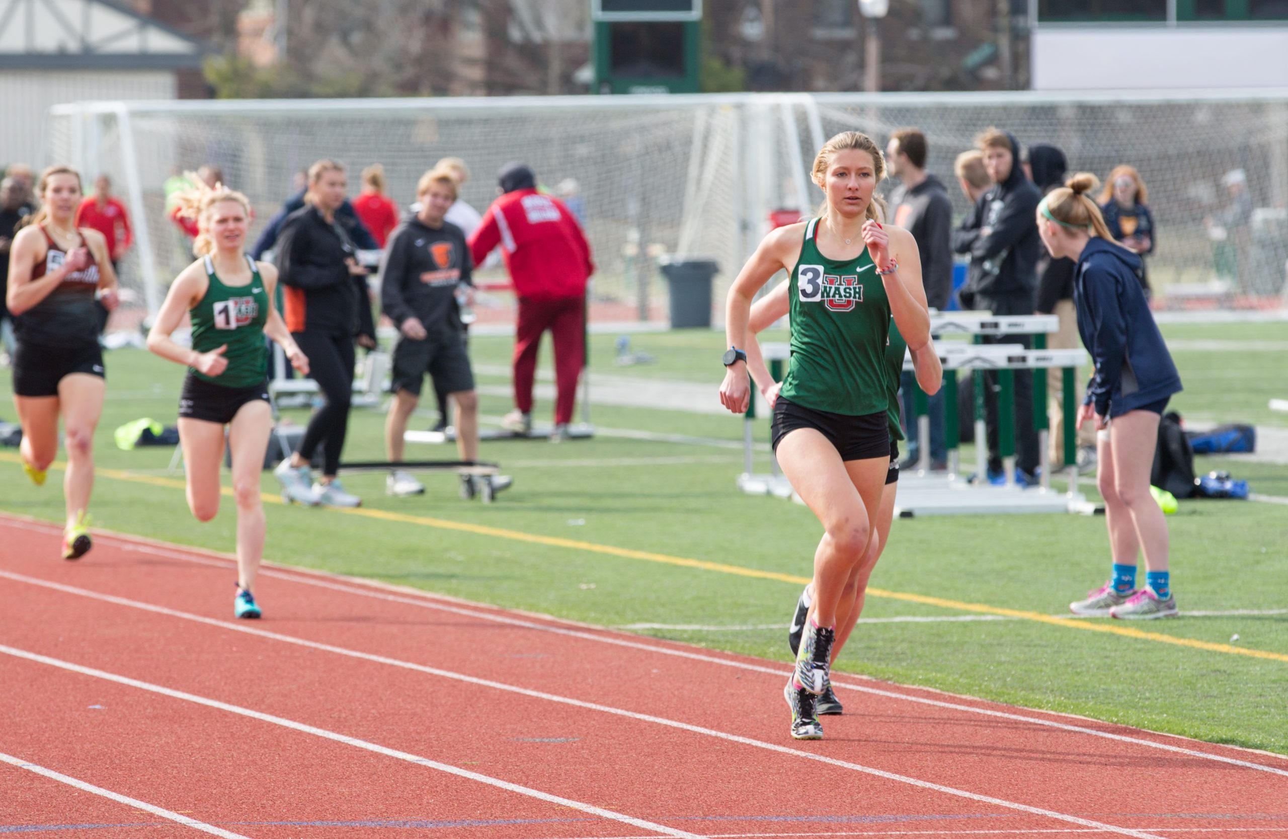 A runner with a green tank top and black shorts strides along the far right side of a track. People mill about in the background.