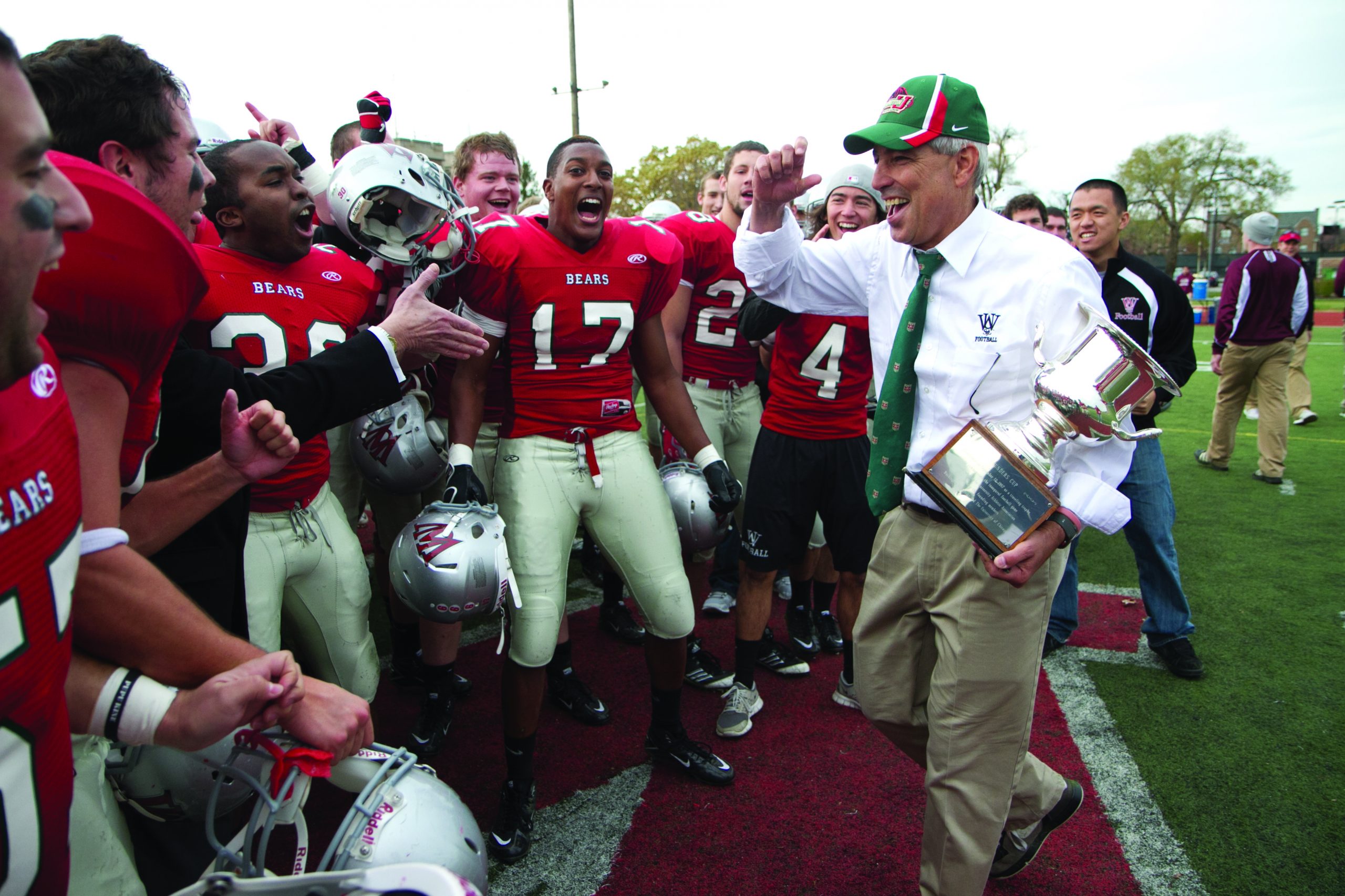 A man in a white button down shirt and green tie holds a trophy in his left hand and raises his right arm toward a green and red cap. He wears beige khakis. To his right are football players in red uniforms and white pants. They are shouting, many with their helmets off.