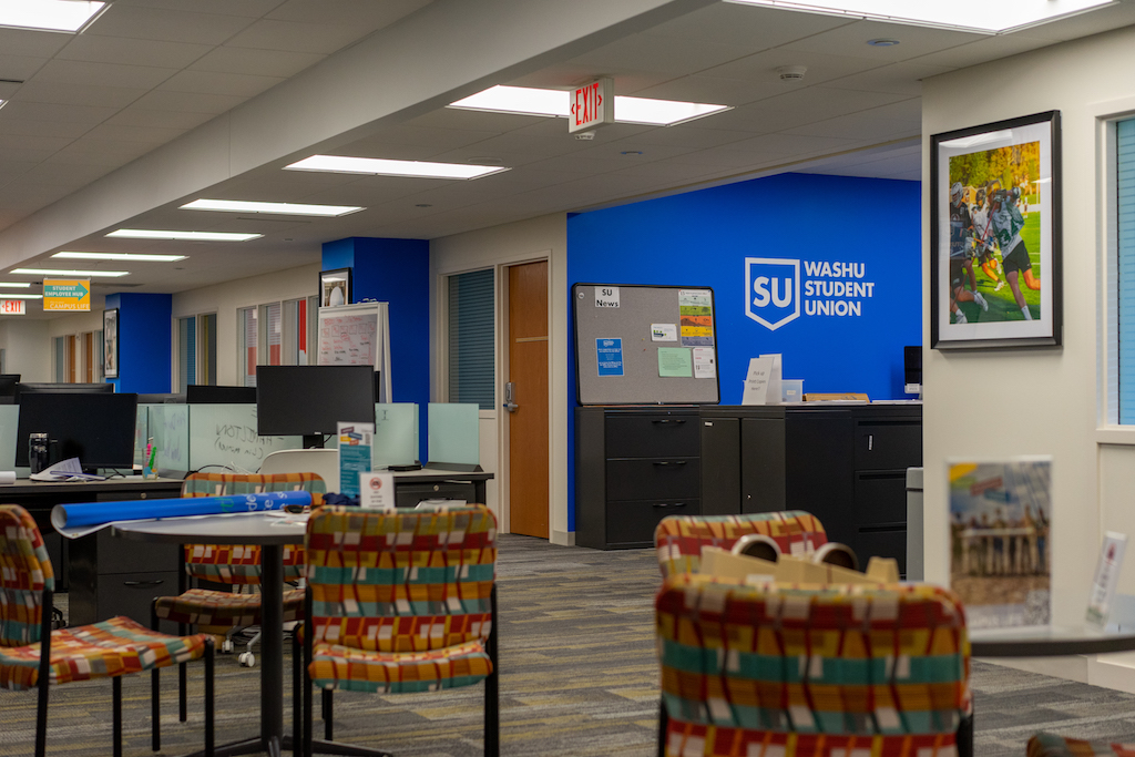 A cluster of patterned chairs stand around a well lit room with a grey carpet. A blue wall will a logo reading "student union" stands in the background next to desks with computers and papers sitting on them