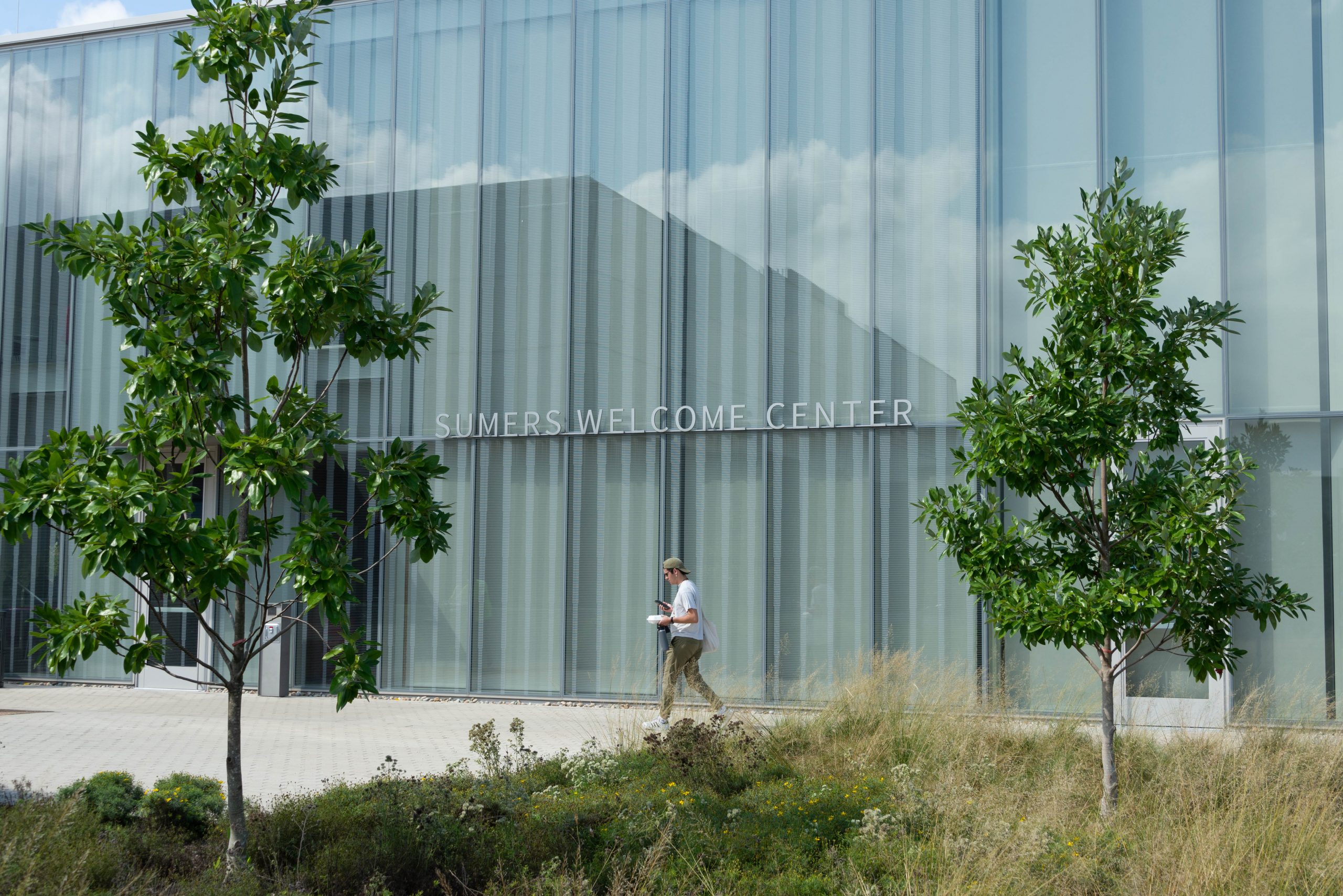 A person in a white shirt walks in front of a glass building framed by green trees.