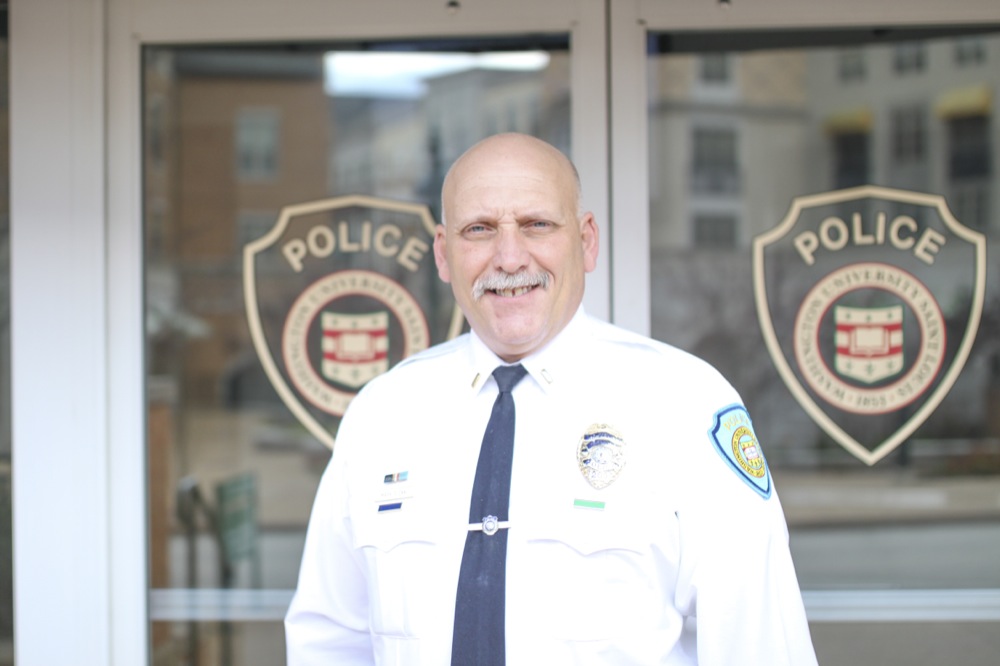 A man with a gray moustache and a bald head stands in front of glass doors with a police insignia on them. He wears a white shirt with a navy blue tie and has a police patch on his left arm.
